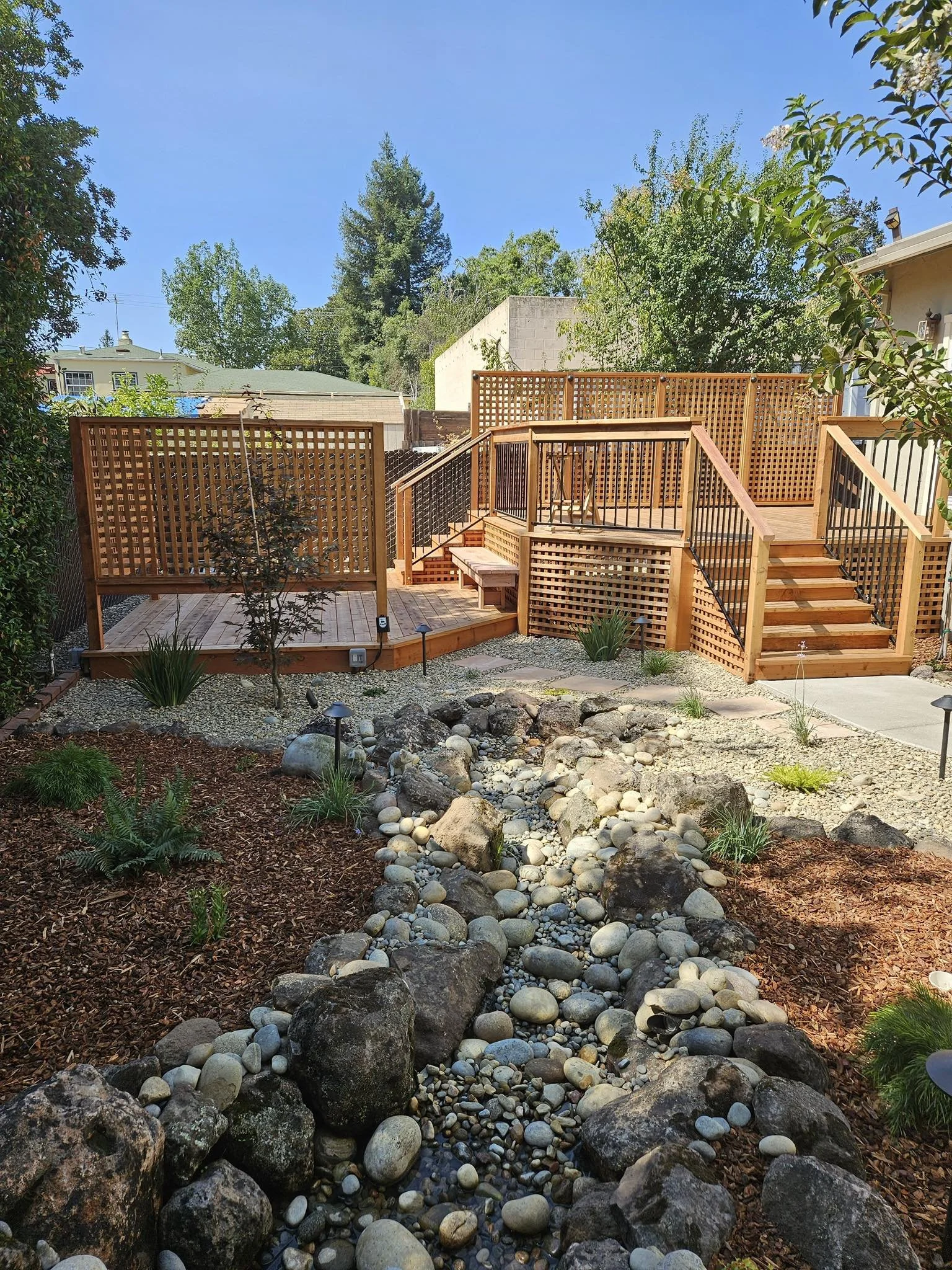 A backyard with a new wooden deck and stairs, surrounded by a garden with trees, shrubs, and a dry creek bed filled with rocks and gravel.
