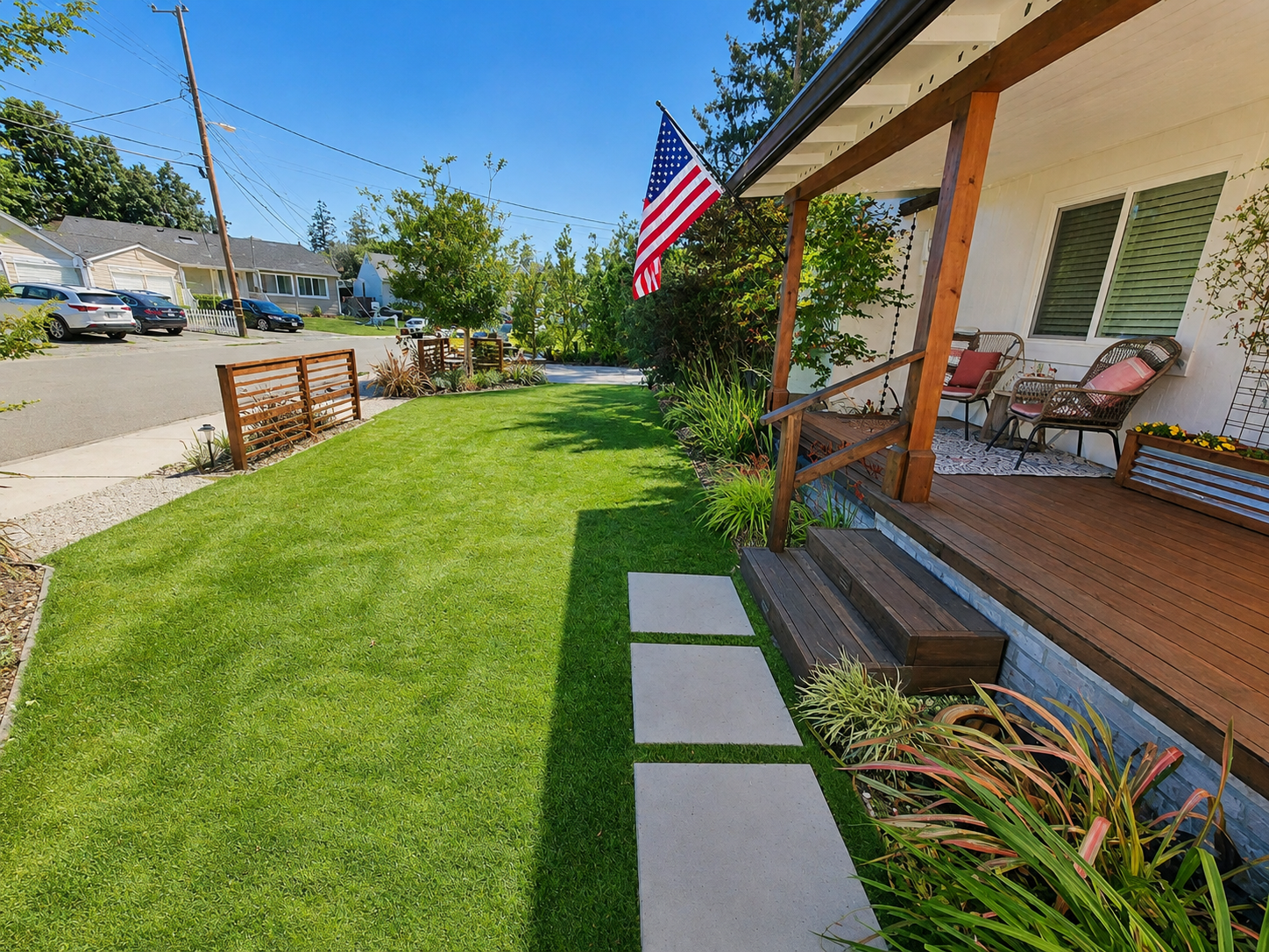 A front yard with a well-maintained green lawn, a wooden porch with chairs and cushions, an American flag hanging from the porch, and a sidewalk with stepping stones. A street with parked cars and houses is visible in the background on a sunny day.