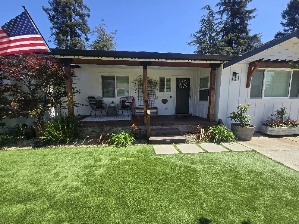 Front view of a suburban house with a small front porch, outdoor chairs, potted plants, a John Deere toy tractor, and an American flag.