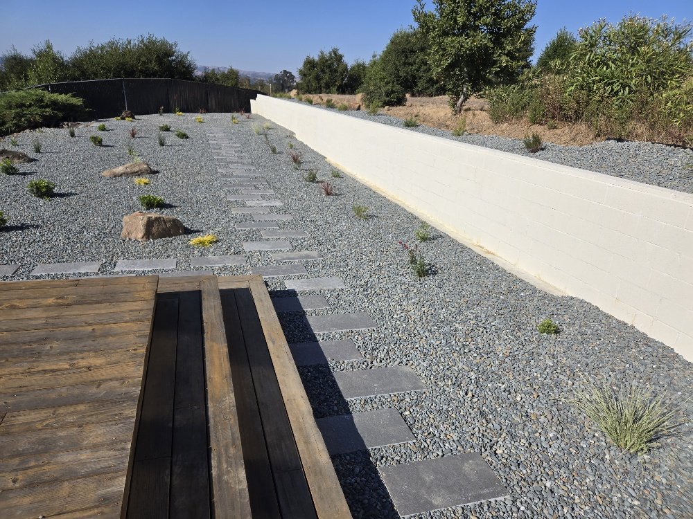 A backyard with a gravel ground, small plants, large rocks, a wooden deck in the foreground, a white brick wall on the right, and bushes and trees in the background under a clear blue sky.