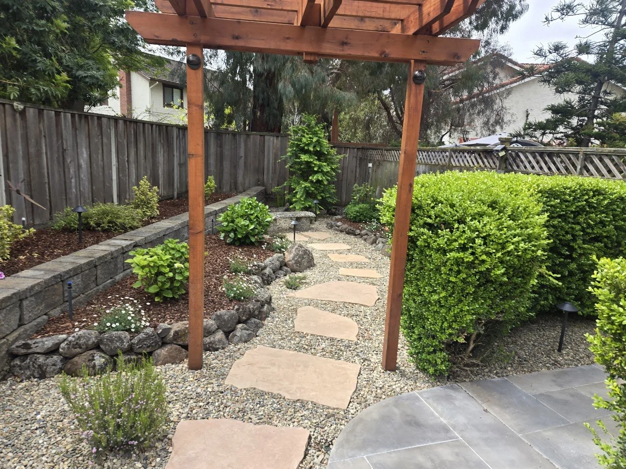 Backyard garden with a gravel pathway, lush green bushes, and a wooden pergola.