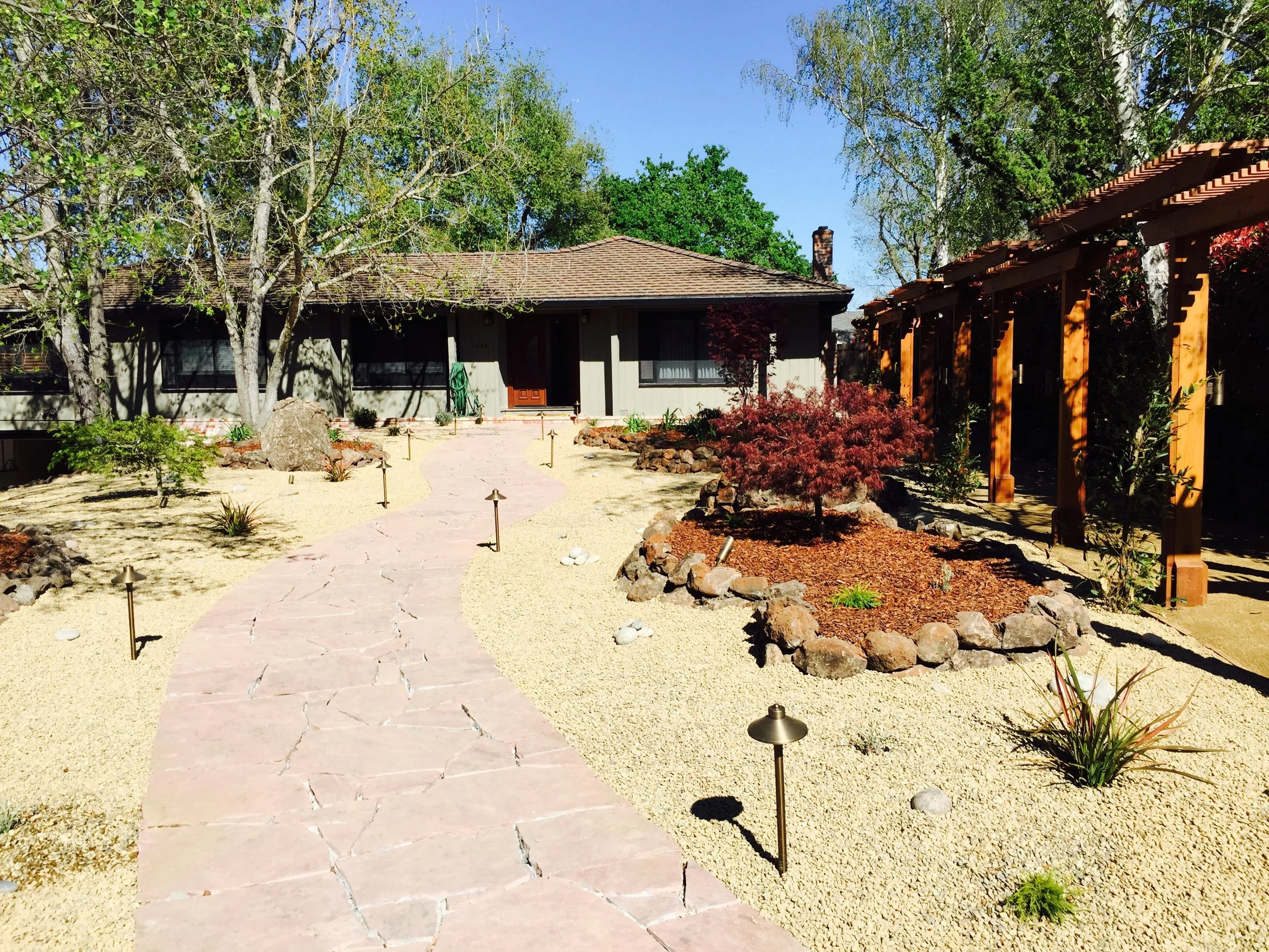 A house with a brown shingled roof, surrounded by a garden with young trees and shrubs. A stone pathway leads to the front door, and there is a wooden pergola on the right side of the garden.