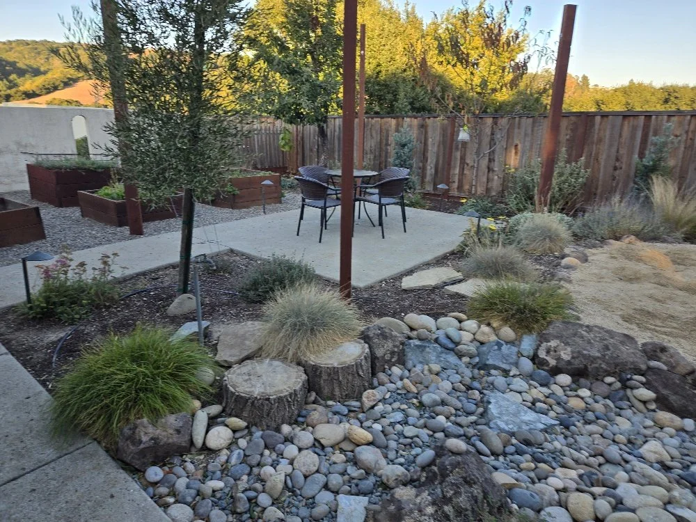 A backyard garden with a small patio and outdoor table with four chairs, surrounding plants, rocks, and a wooden fence in the background.