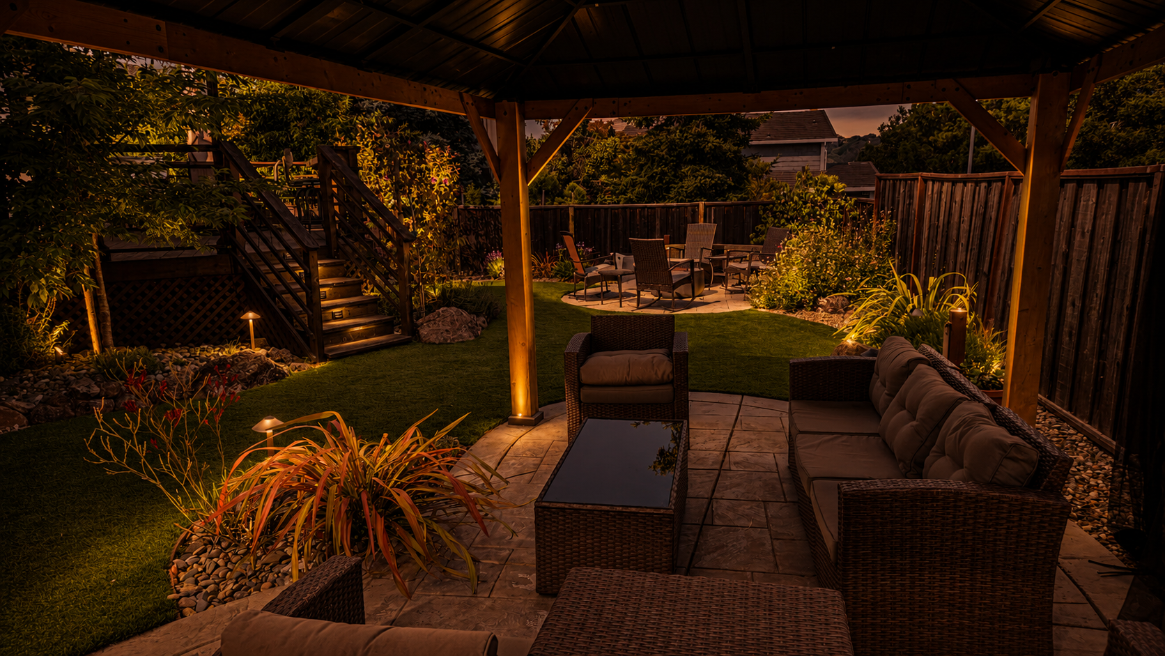 Illuminated backyard patio area at dusk with wicker furniture, a fire pit, a lawn, and a raised seating area, enclosed by a wooden fence and surrounded by trees and shrubs.