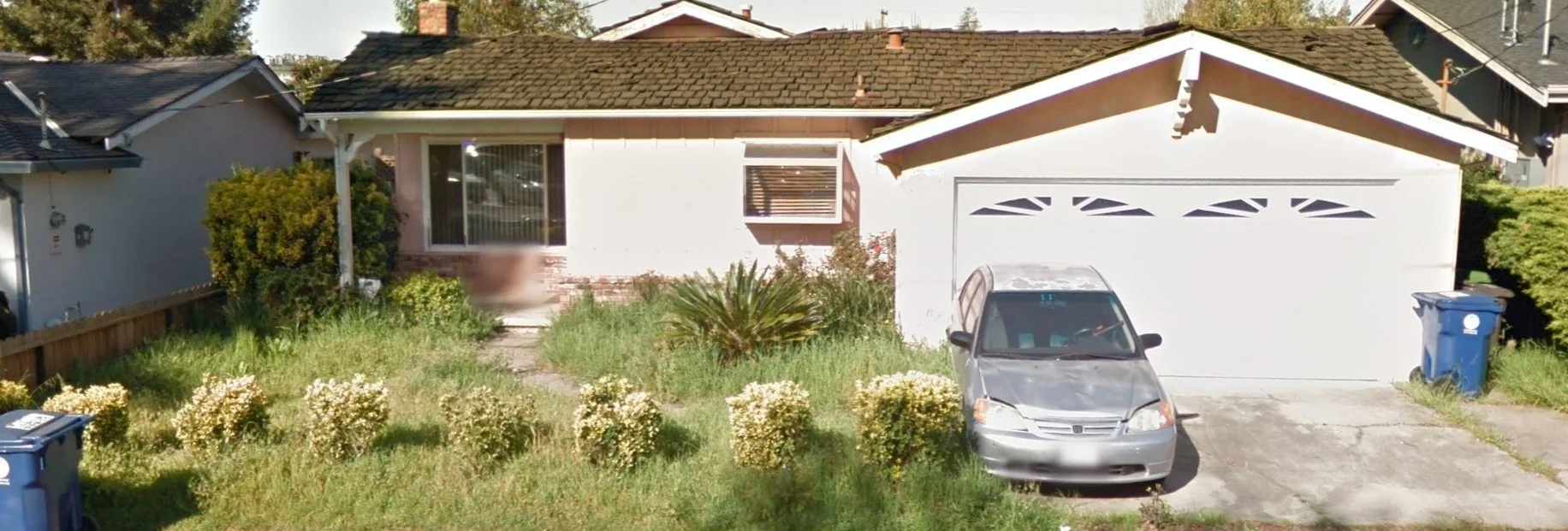 Front view of a single-story house with a white exterior and a small front yard overgrown with grass and bushes, a silver car parked in the driveway, and blue trash bins on the sides.