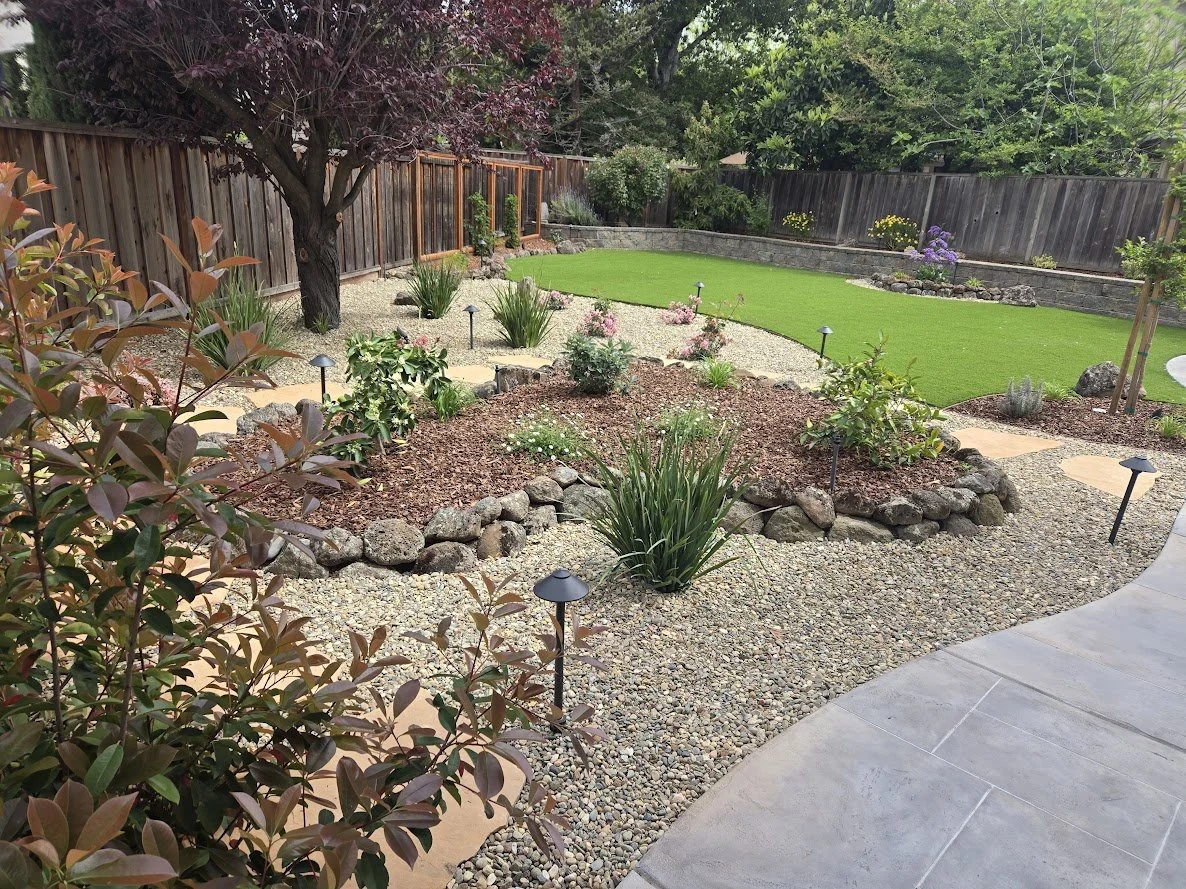 A landscaped backyard garden with a curved concrete patio, gravel pathways, flower beds, a large tree, and a wooden fence. The garden features green grass, plants, flowers, and landscape lighting.