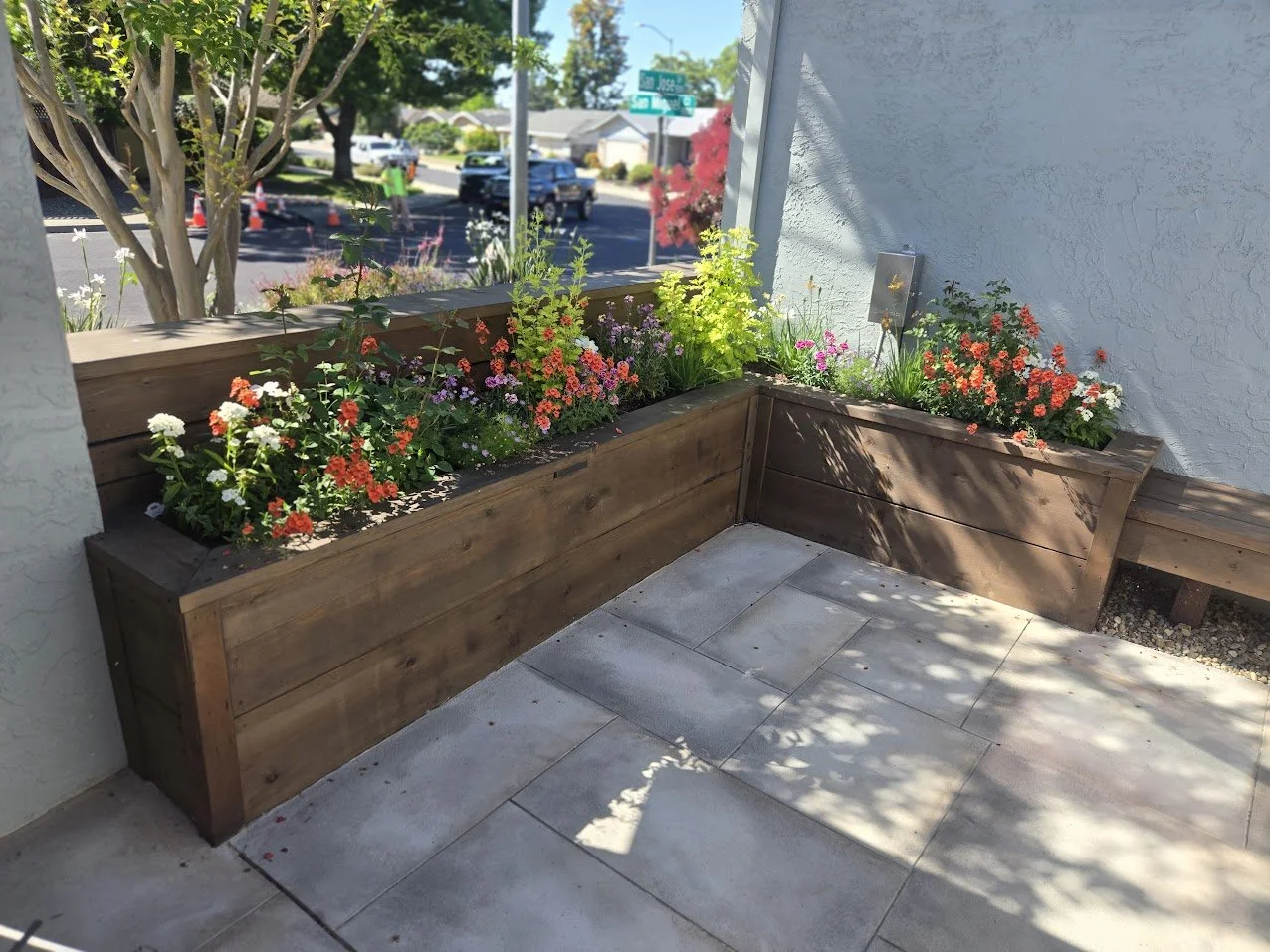 Flower bed with colorful flowers in wooden planter boxes outside a building on a sidewalk, with a street and cars passing by in the background.