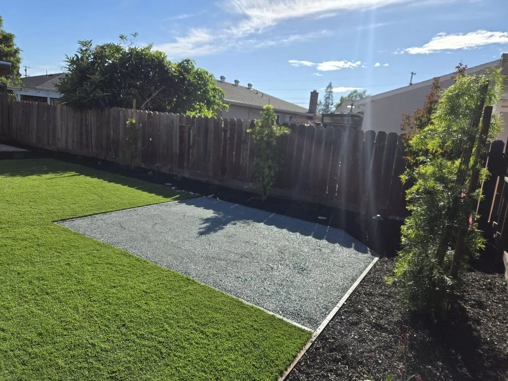 Backyard with well-maintained green grass, a gravel patch, wooden fence, small trees, and shrubs under a sunny sky.