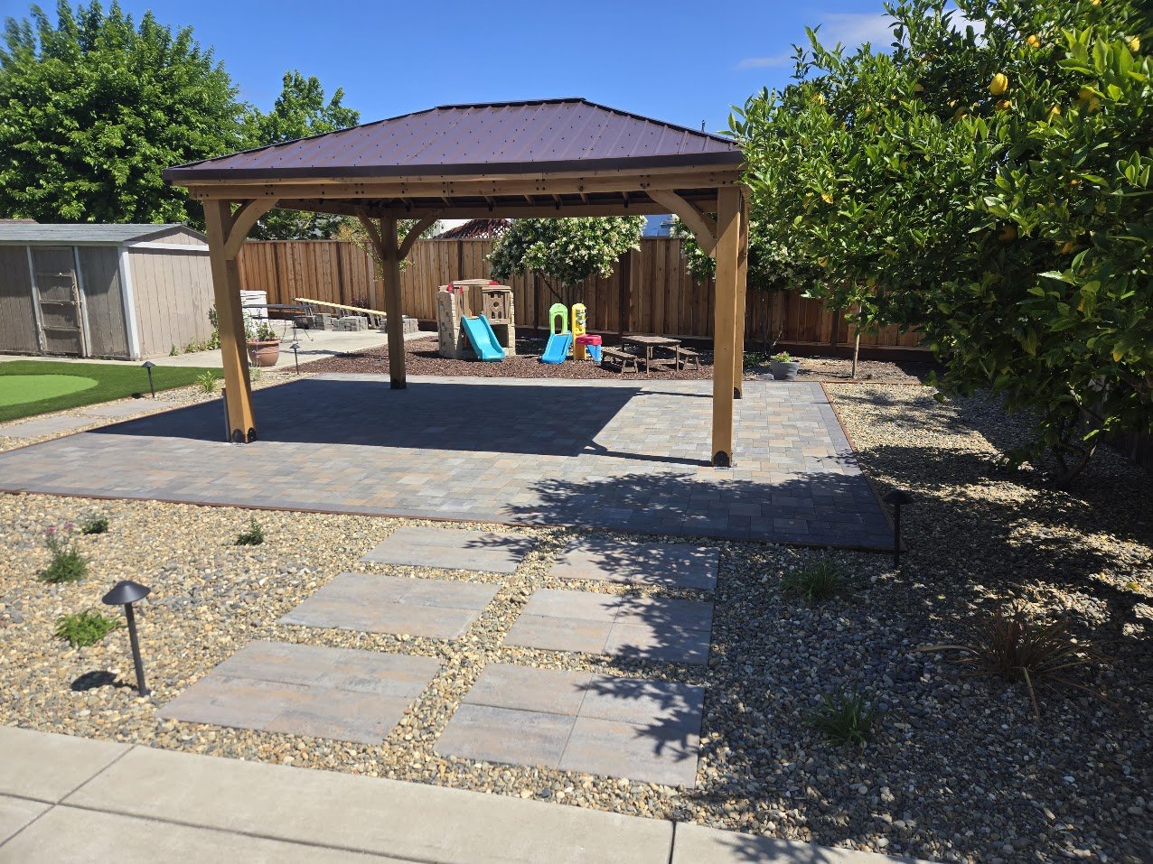 Backyard with a covered pavilion, paved patio, small playground, and landscaped garden.