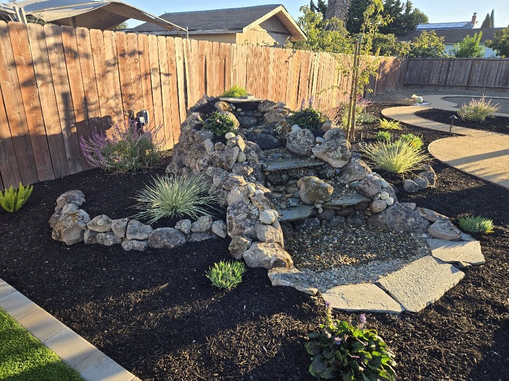 Backyard garden with a small stone waterfall feature surrounded by various plants and mulch, enclosed by a wooden fence.