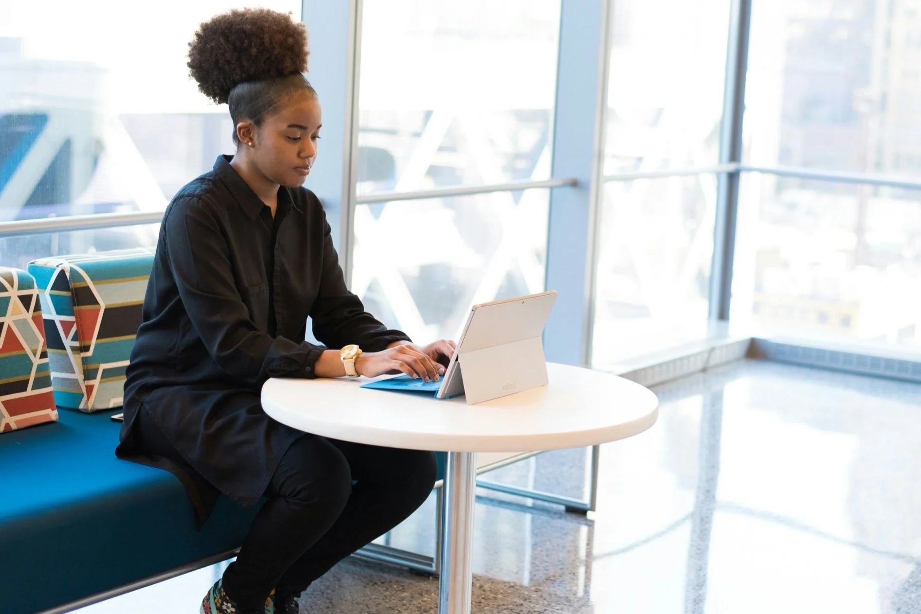 A young woman with curly hair in a high puff hairstyle, wearing a black shirt and a watch, sitting at a white round table in a modern building with large windows, using a tablet. There are patterned bags on a blue bench beside her.