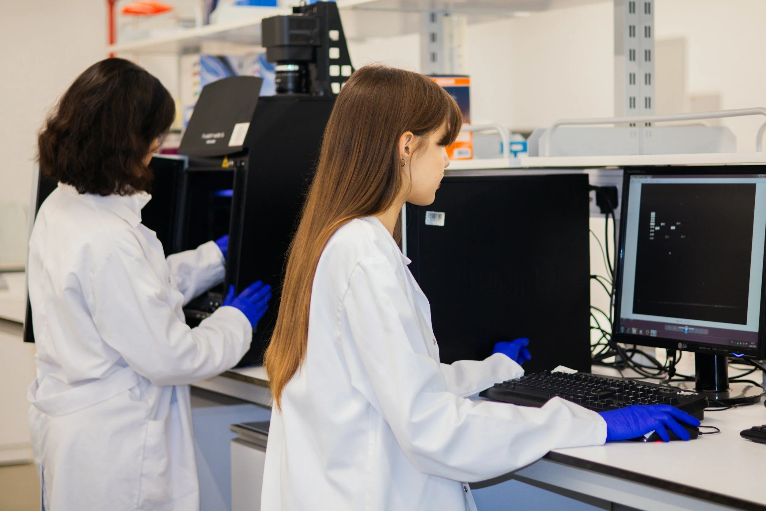 Two scientists in white lab coats and blue gloves working in a laboratory, using computer monitors and scientific equipment.