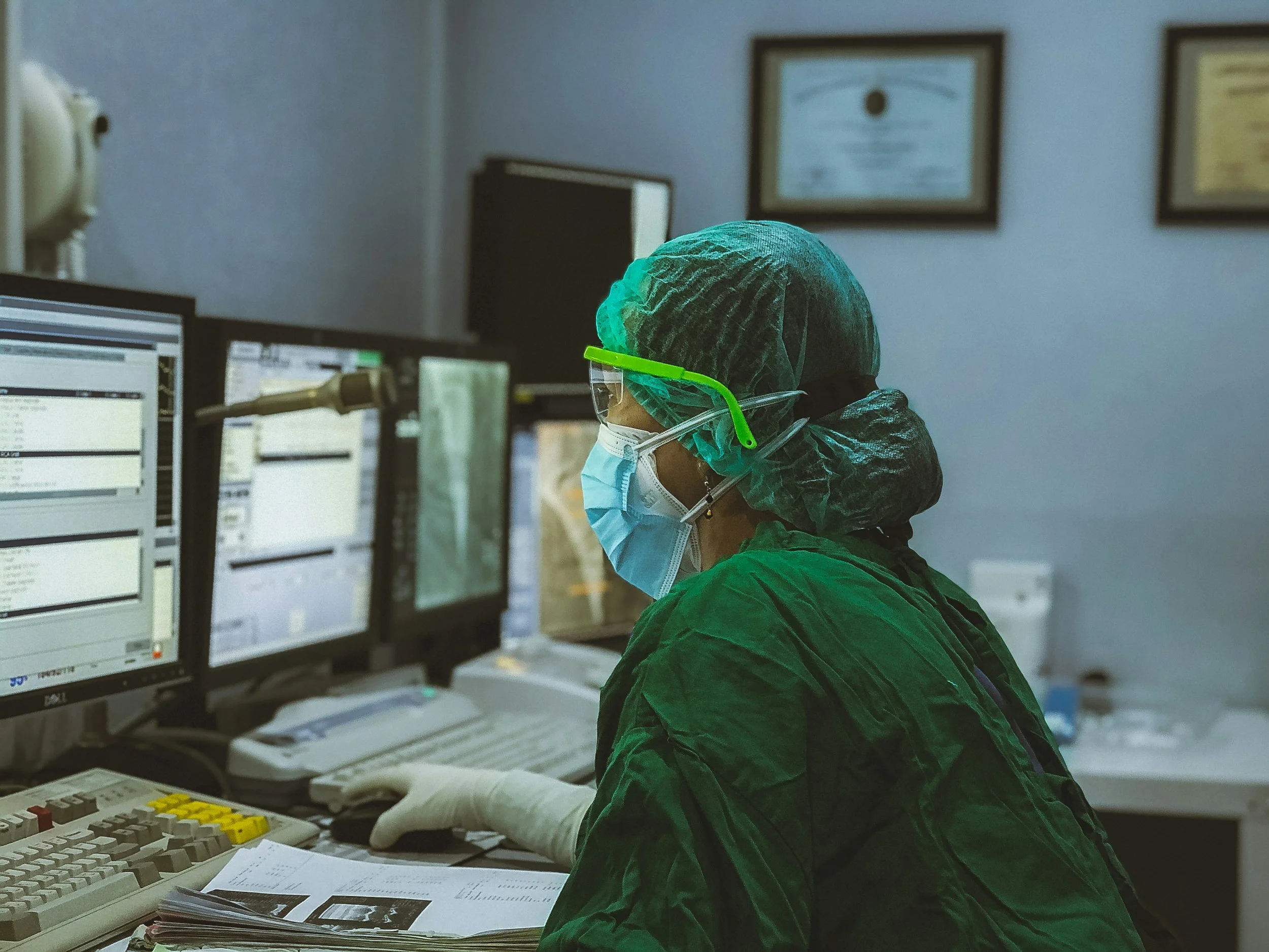 A healthcare worker wearing a green protective gown, hair cover, face mask, and safety glasses is working at a computer in a medical office or hospital. The worker is looking at a monitor with lab results or medical data, and there are multiple computer screens, a telephone, and medical charts on the desk.