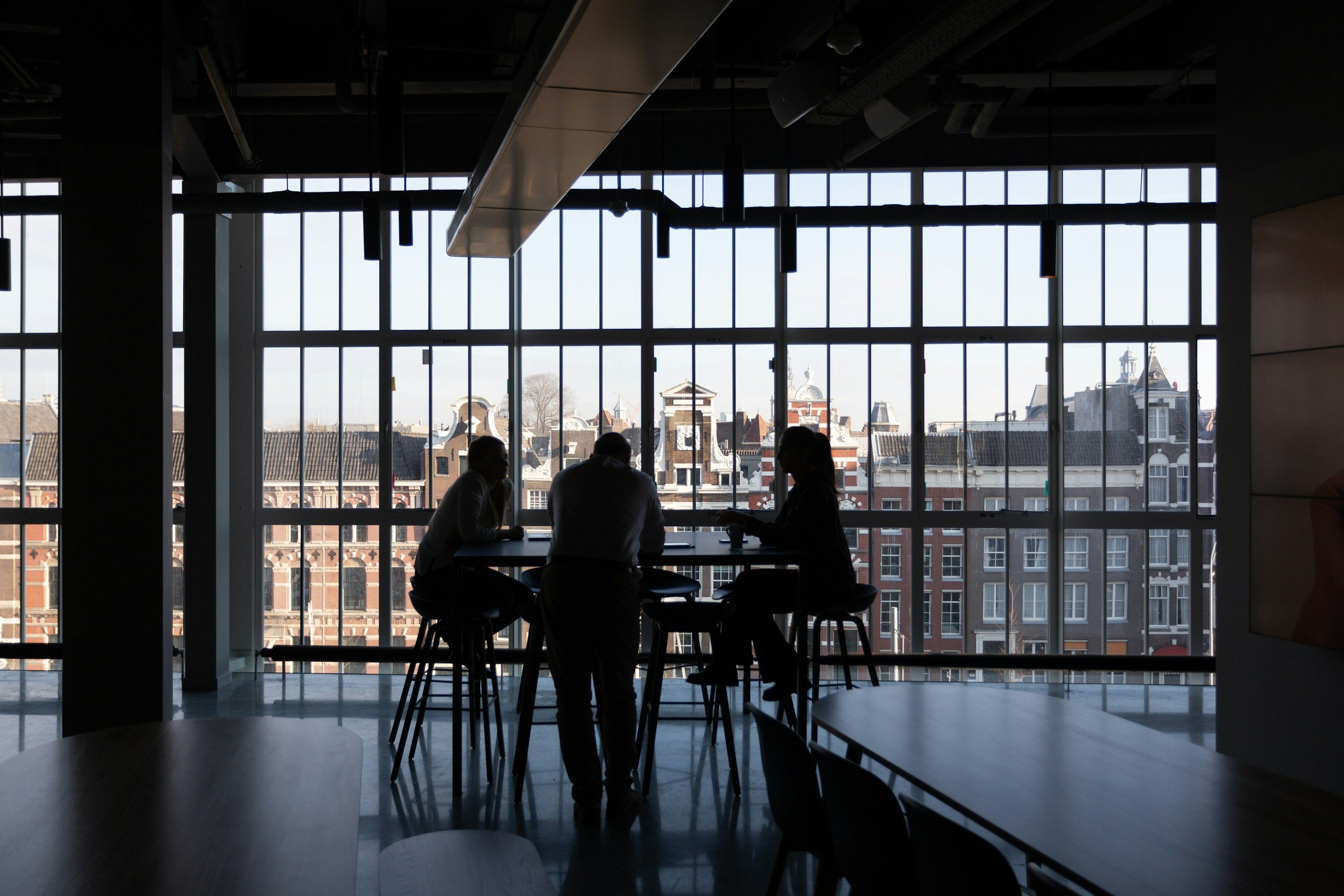 Silhouettes of three people sitting at a table inside a modern building, with large windows revealing a cityscape of historic buildings and a clear sky outside.