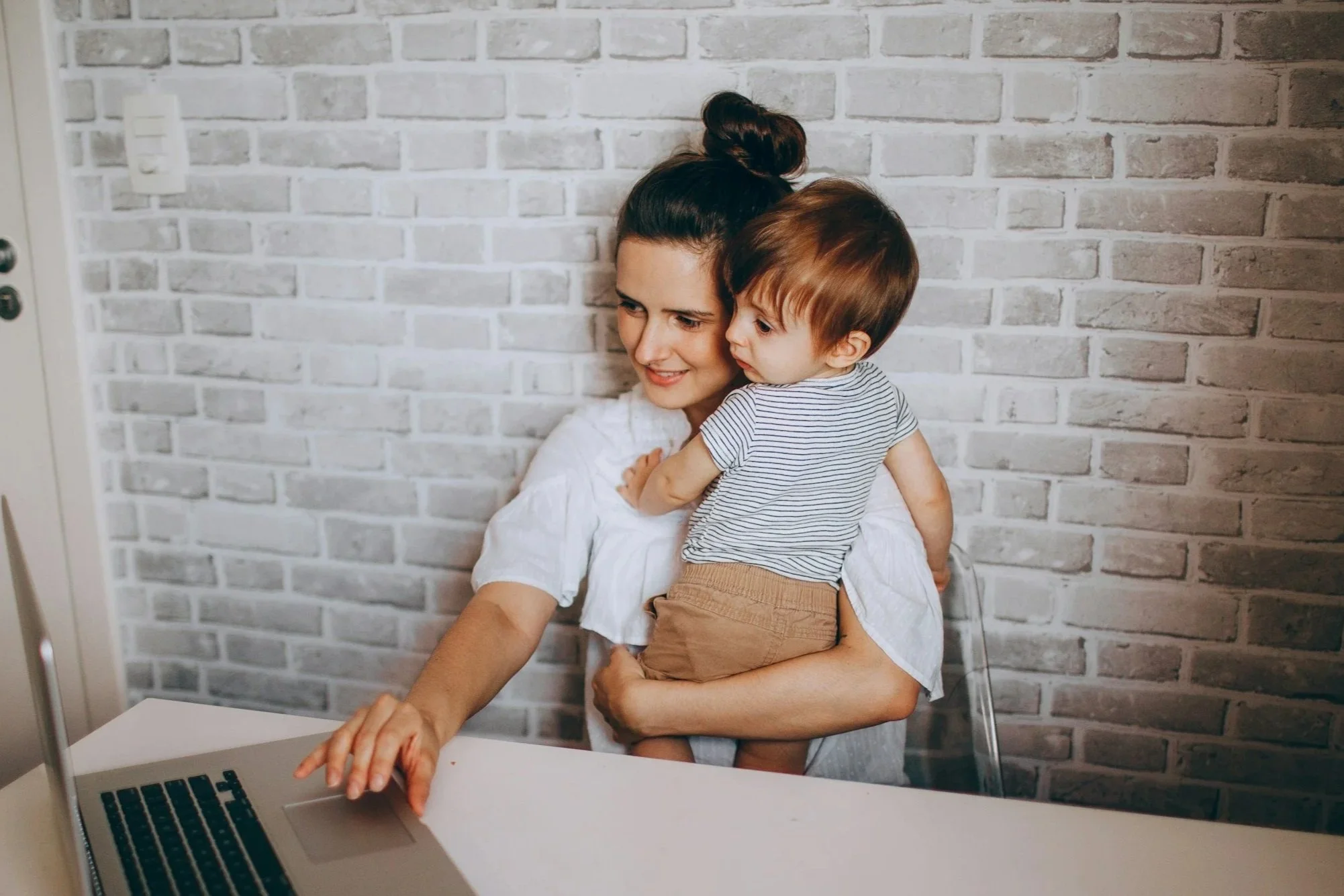A woman with dark hair in a bun holding a young boy with brown hair in front of a white brick wall, looking at a laptop on a white table.