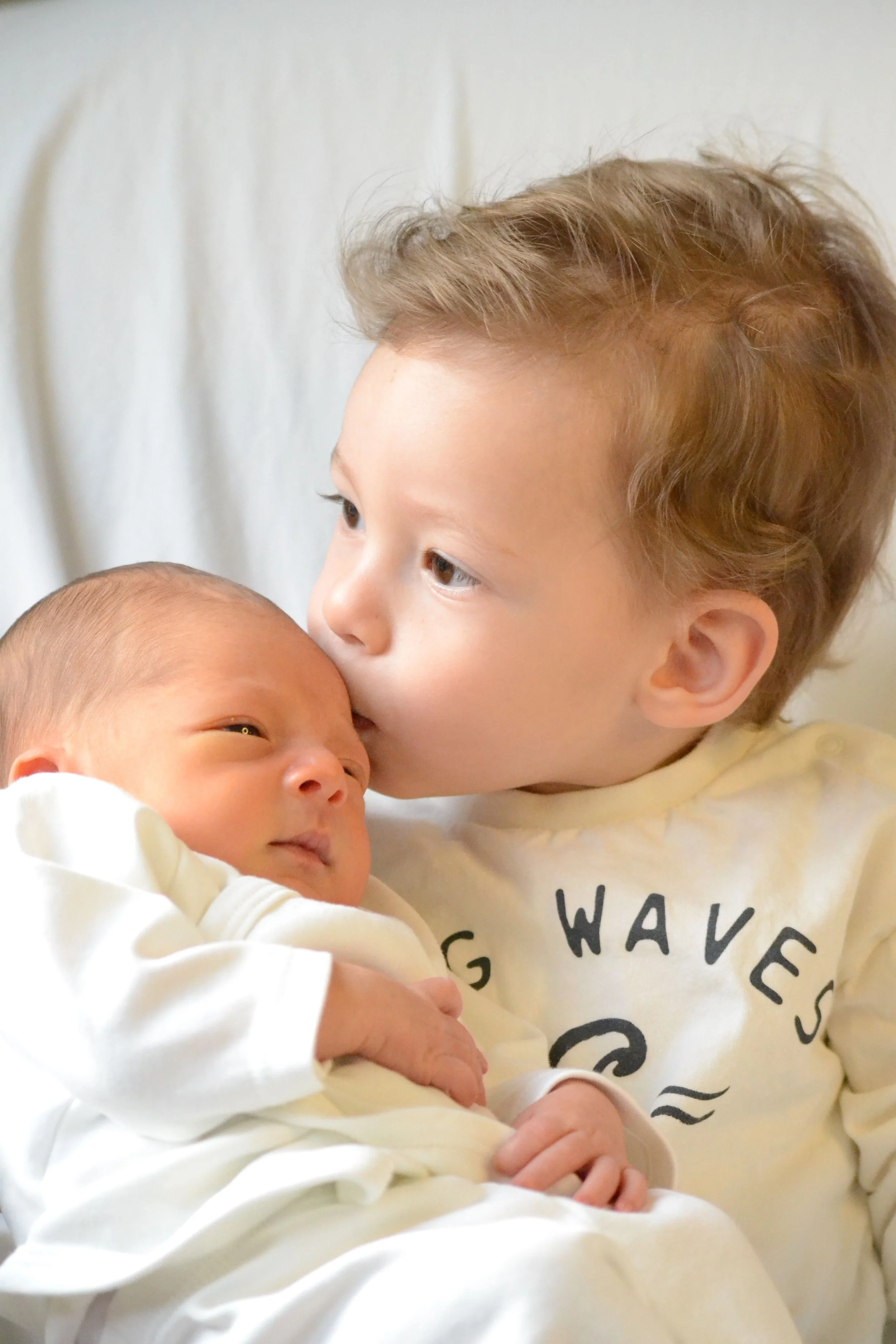 A young boy holding a newborn baby, gently pressing his face against the baby's head, both lying on a white surface, with the boy wearing a light-colored shirt with text.