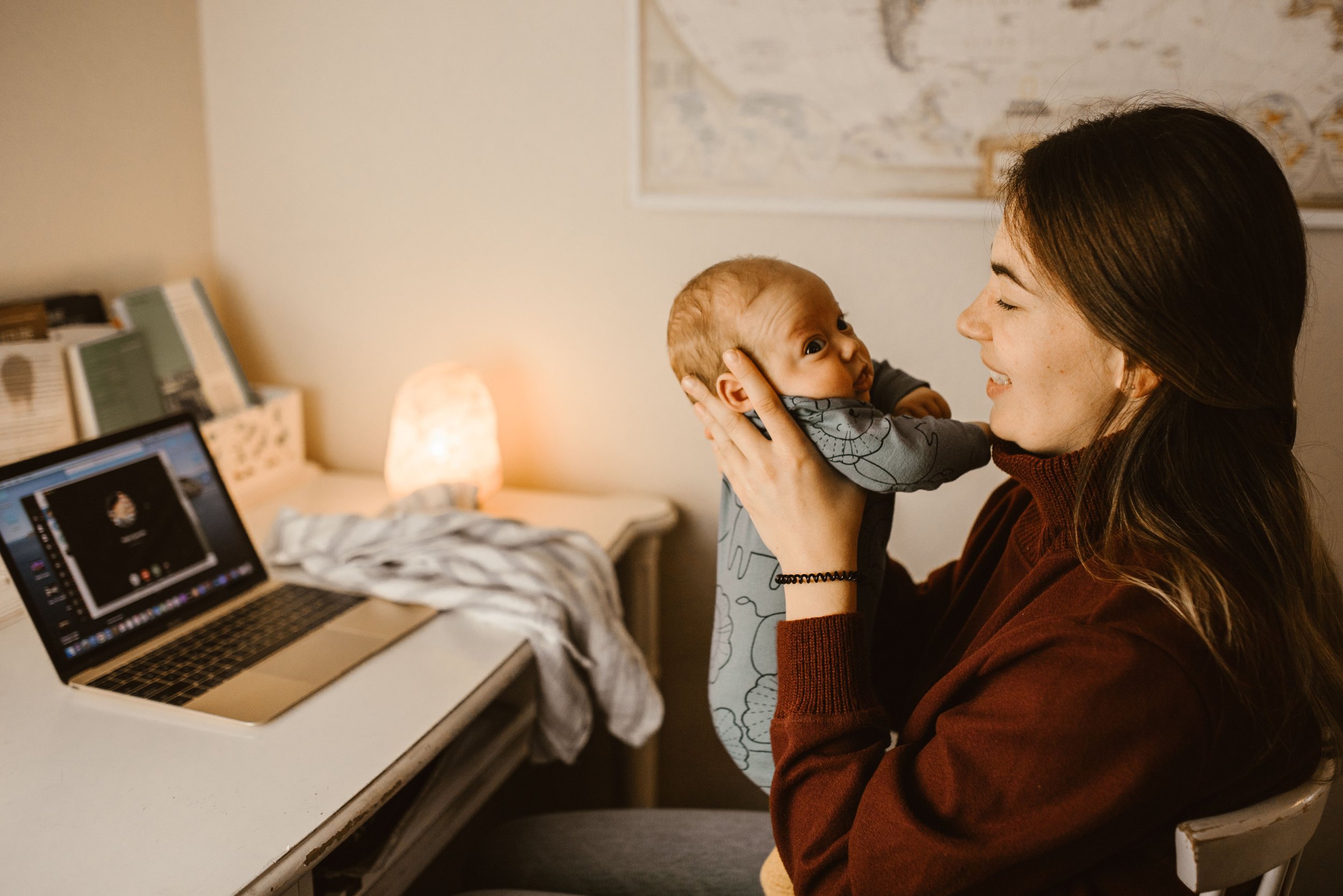 A woman holding a baby face-to-face in a warmly lit room with a desk, laptop, and books in the background.