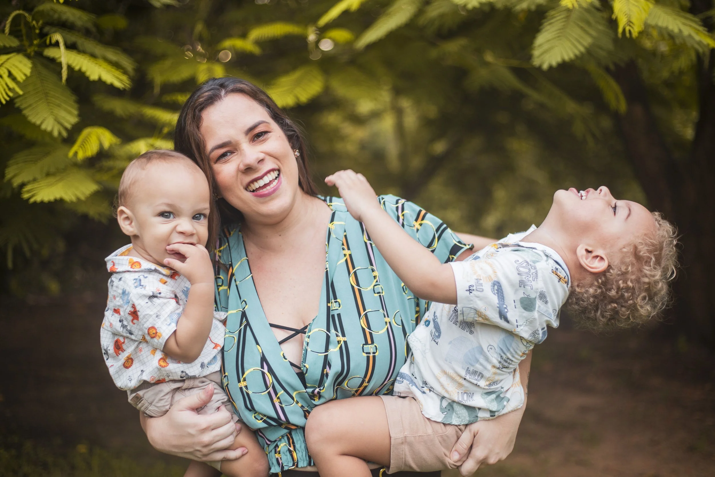 A woman holding two children, one on each arm, outdoors with green foliage in the background. The children are smiling and laughing, and the woman has a big smile on her face.