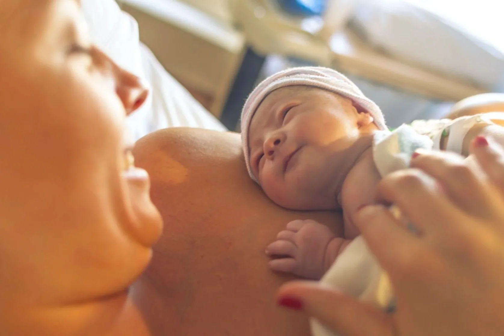 A newborn baby is breastfeeding, held by a woman, possibly the mother, with another person, likely a sibling, looking on. The baby is wearing a pink striped hat and appears happy. The scene takes place in a hospital or medical setting.
