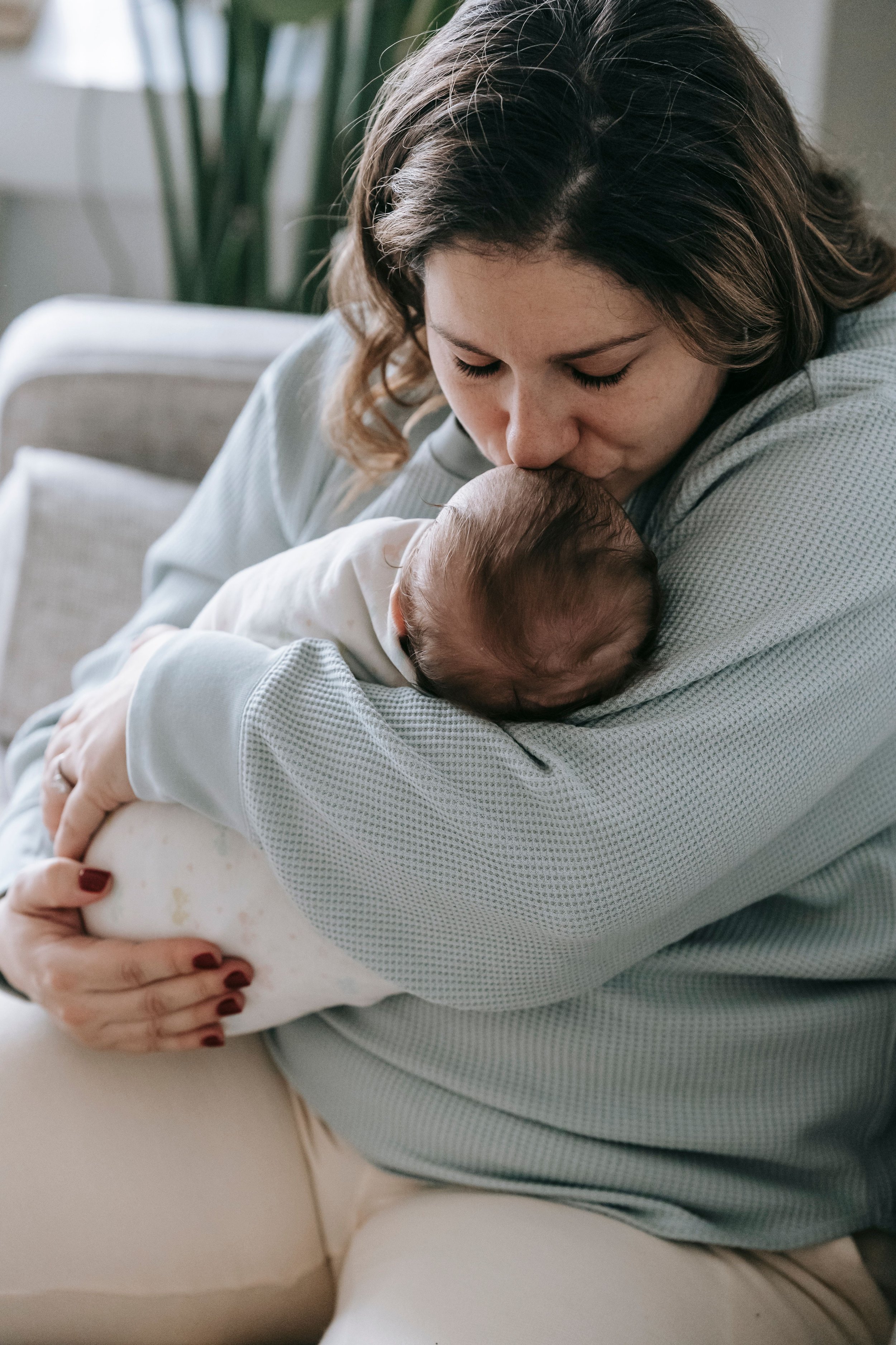 A woman holding a baby close for a kiss in a cozy indoor setting with a sofa and a plant in the background.