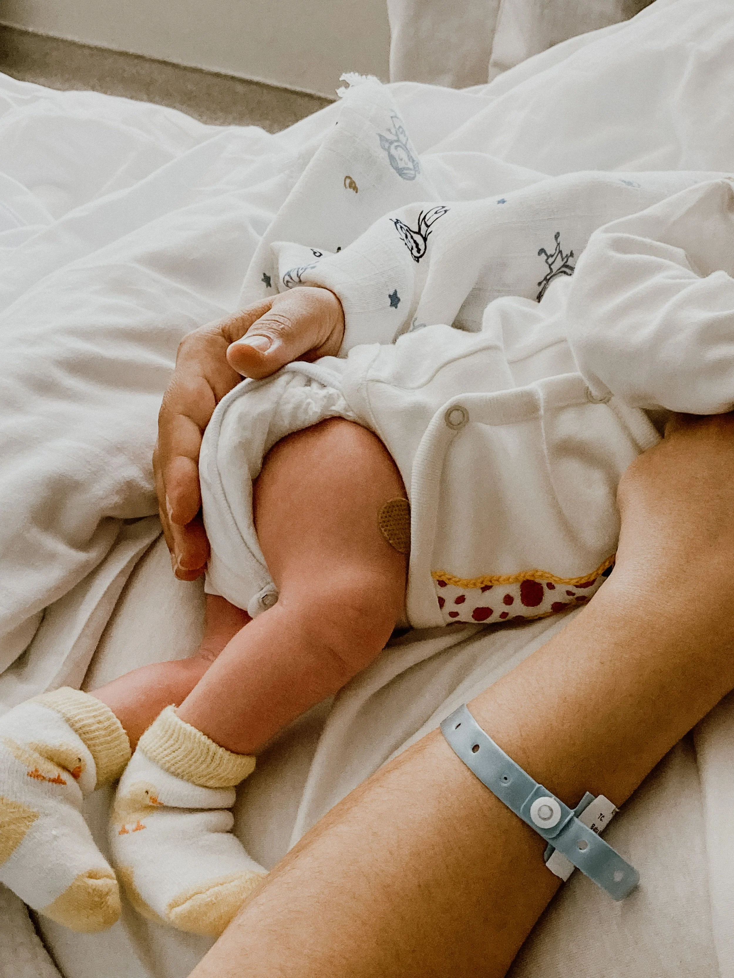 Close-up of a newborn baby's hand holding an adult's finger, with hospital wristbands and blanket visible.