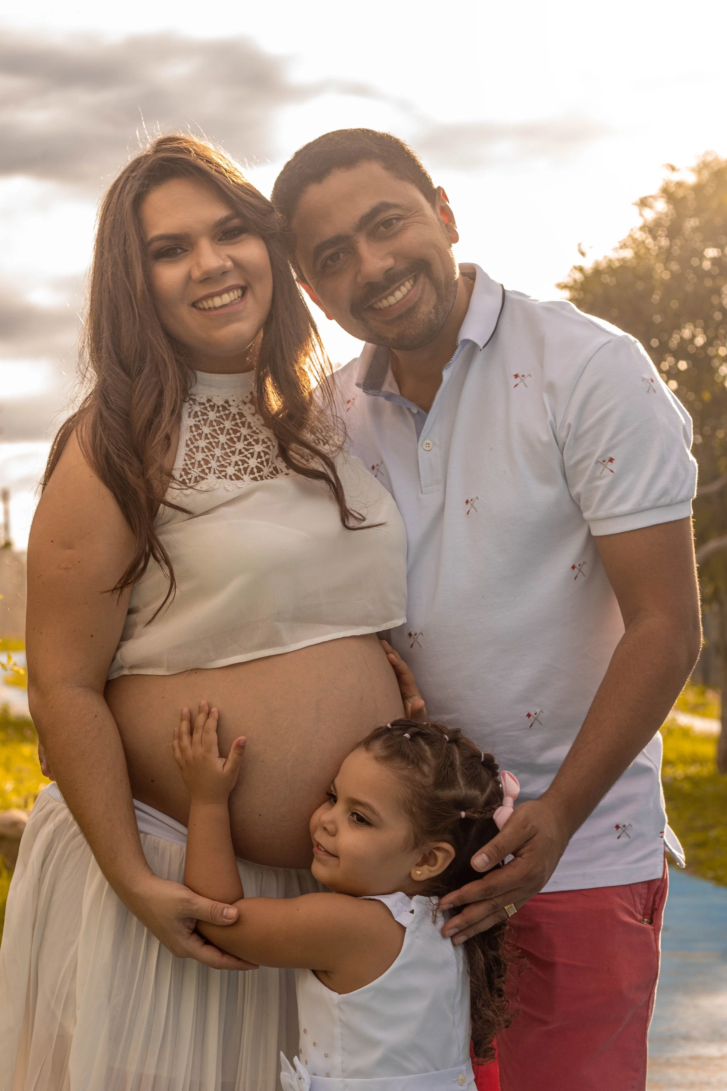 A pregnant woman in a white dress with lace detail on the chest, standing outdoors during sunset, smiling while her young daughter with curly hair and a white dress hugs her tummy, and an adult man in a white polo shirt with small red crossed tools pattern, and red pants, smiling and standing behind her in a park by a body of water.