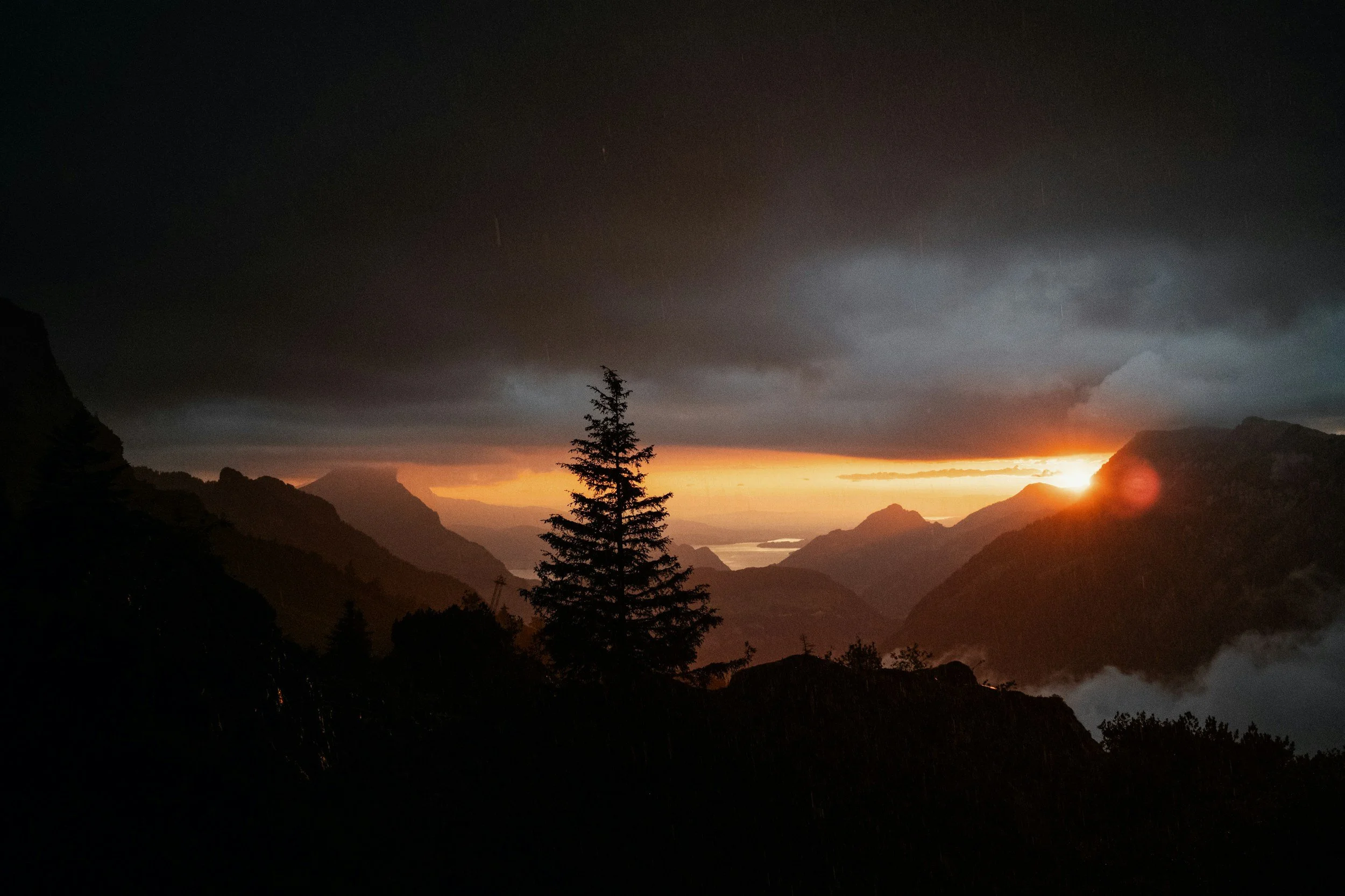 Mountain landscape during sunset with dark clouds, a single pine tree in the foreground, and rays of sunlight breaking through clouds over distant mountains.