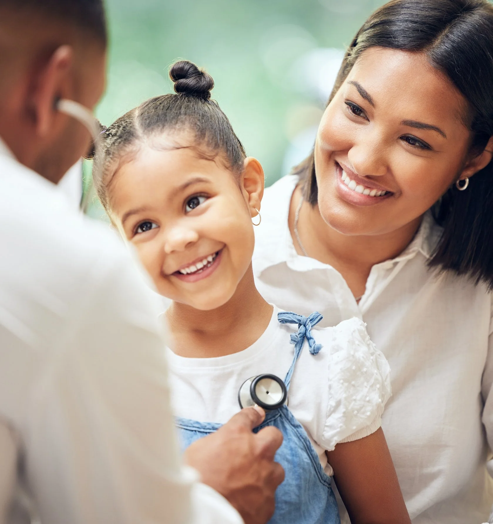 A young girl with a big smile getting her medical checkup from a healthcare professional, with a smiling woman possibly her mother, in a medical setting.