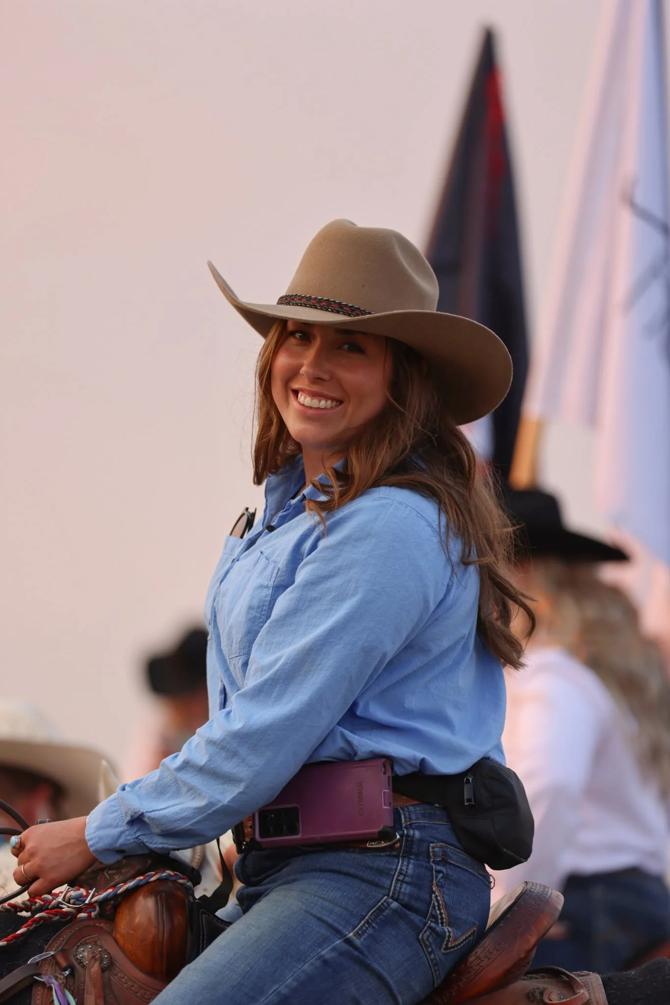 A woman smiling while riding a horse at an event, wearing a cowboy hat, a blue shirt, and jeans, with a smartphone in a belt pouch.