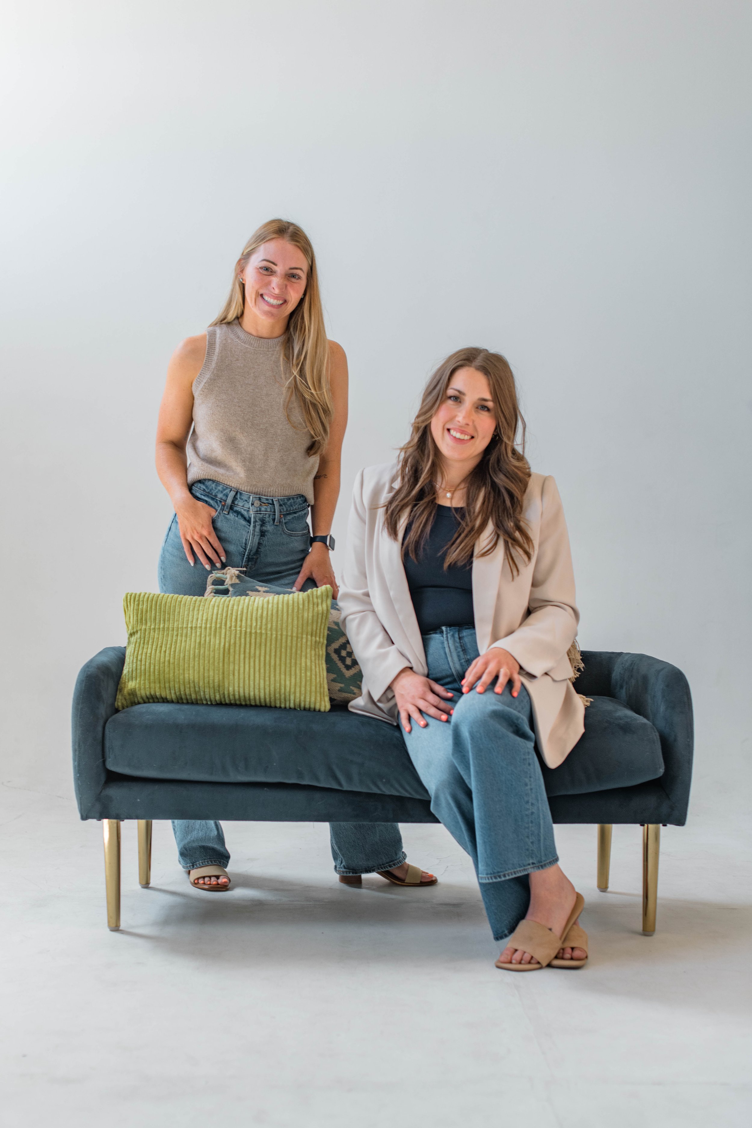 Two women with long hair, one standing and one sitting on a dark blue sofa, smiling at the camera. The standing woman is in a sleeveless beige top and jeans, the seated woman in a light blazer and jeans. A green cushion is on the sofa.