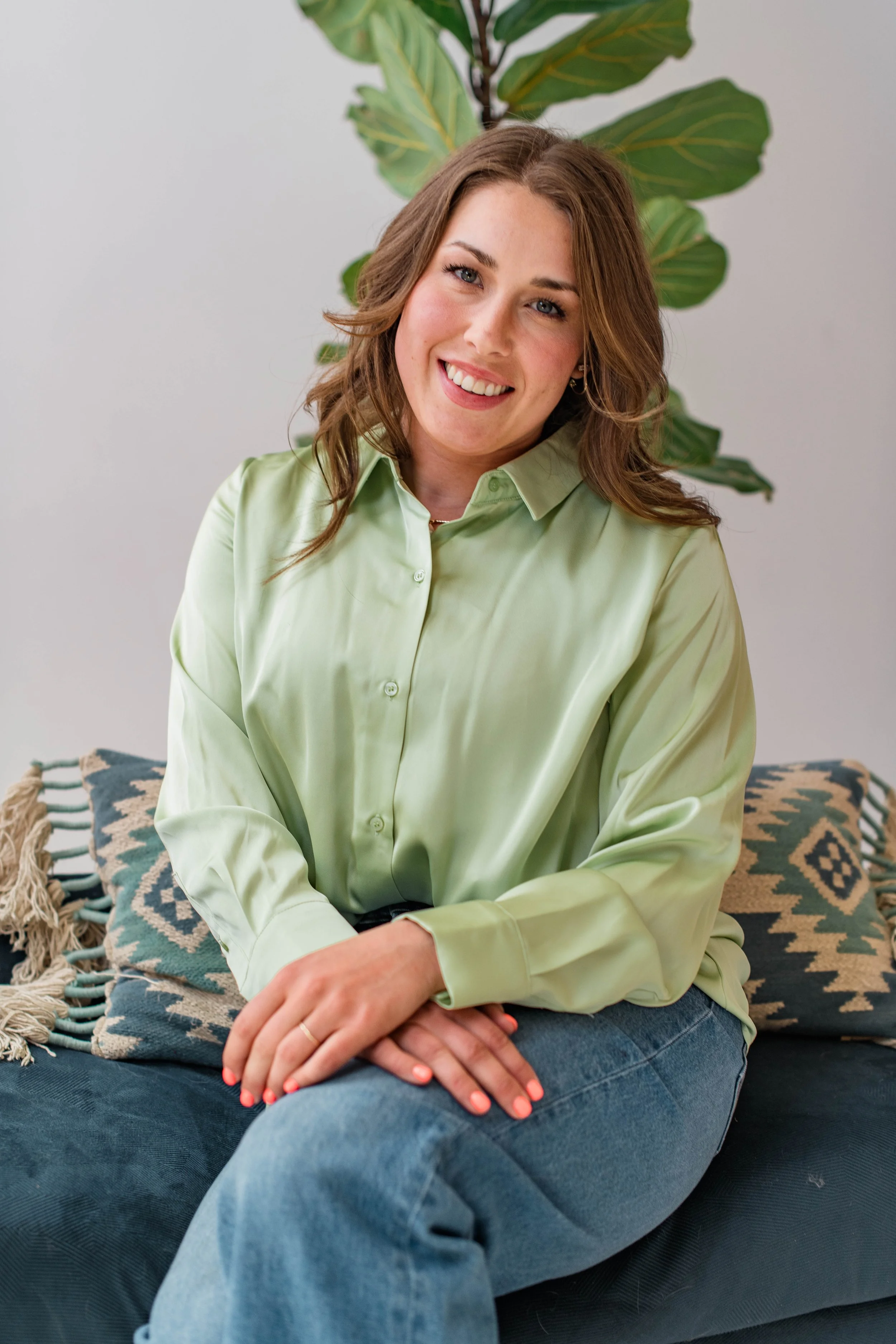 A woman with wavy brown hair and blue eyes is sitting on a dark sofa with patterned pillows. She is wearing a light green button-up shirt and blue jeans, with her hands lightly clasped on her lap. A large green leafy plant is in the background, and her face is smiling gently at the camera.