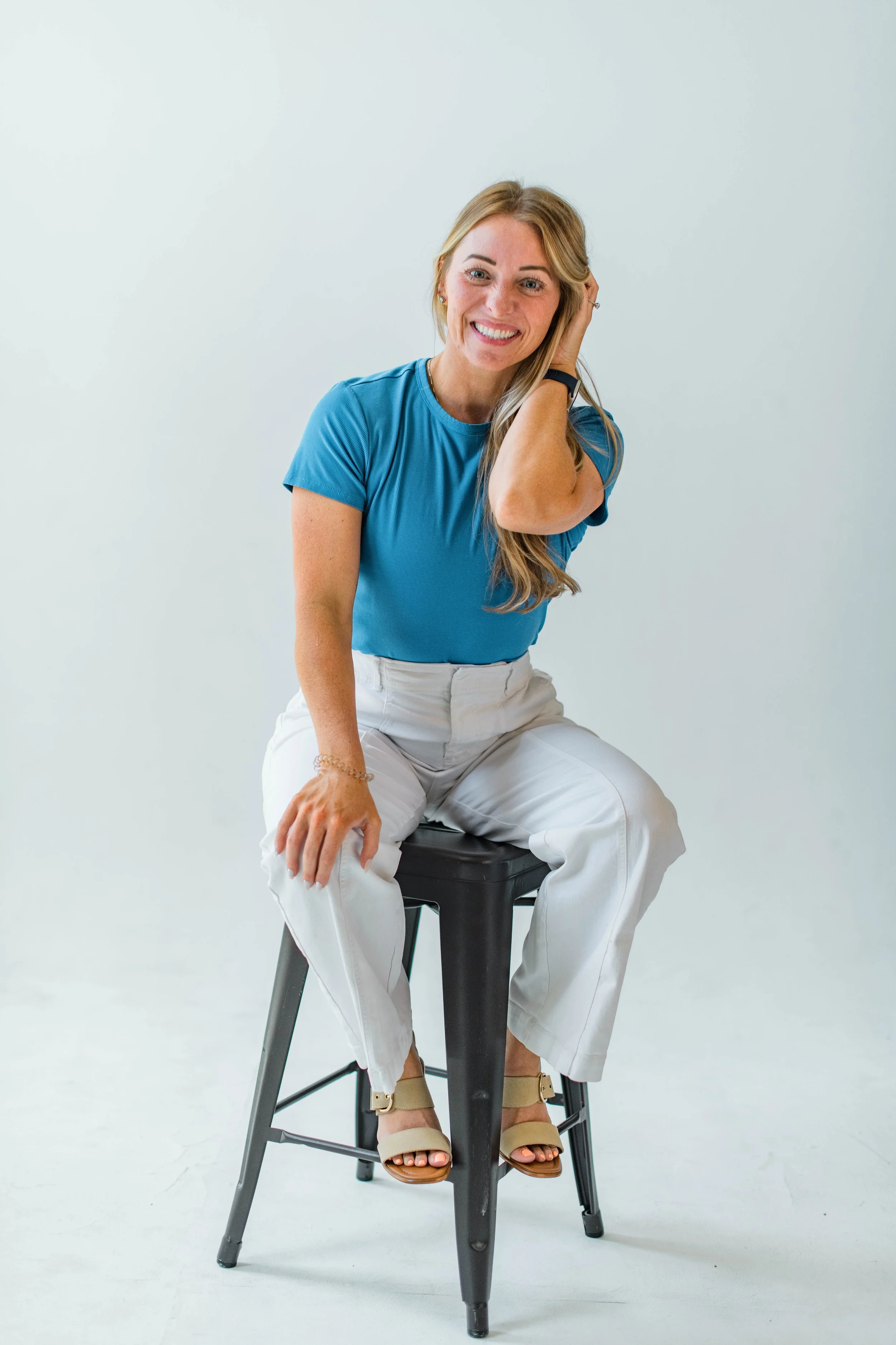 A woman with blonde hair, wearing a blue t-shirt and white pants, is sitting on a black stool in front of a plain white background. She is smiling, with her right hand resting on her head and her left hand on her knee, wearing beige sandals.