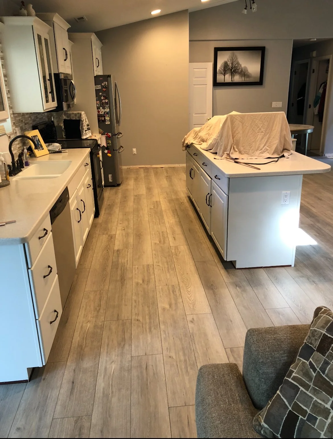 A kitchen with white cabinets, a white island covered with cloth, and wood-look flooring. The refrigerator has magnets, and there's a black sofa with patterned pillows in the foreground.