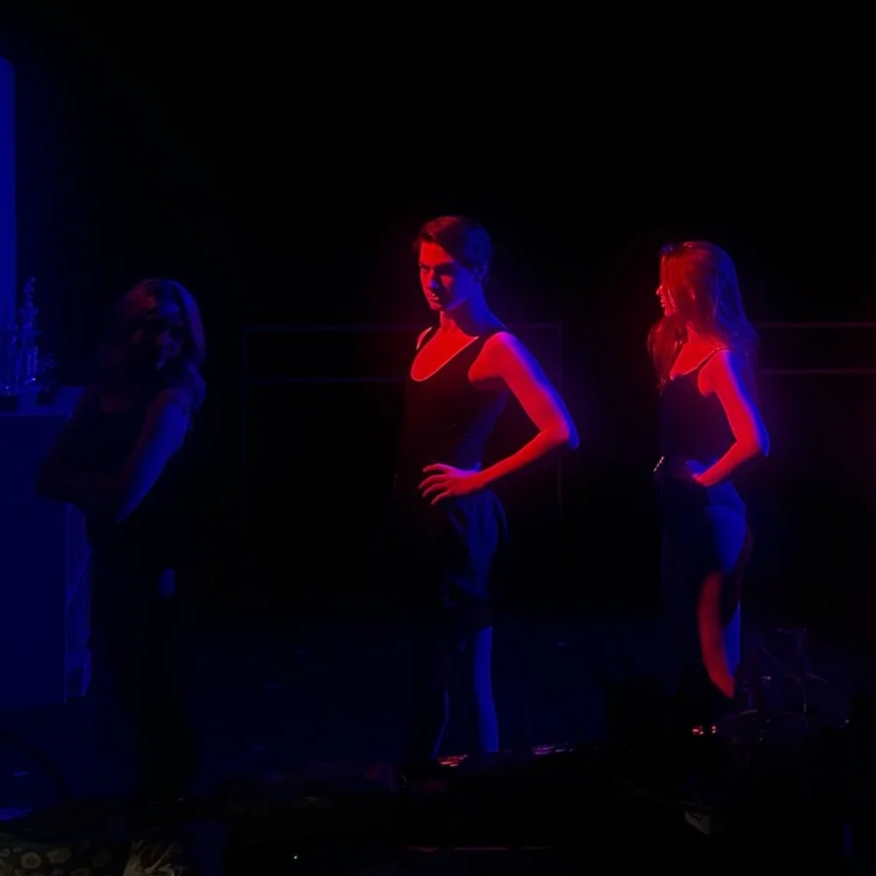 Three women standing in a dark room illuminated by red and blue neon lights, wearing black outfits, posing with confident stances.