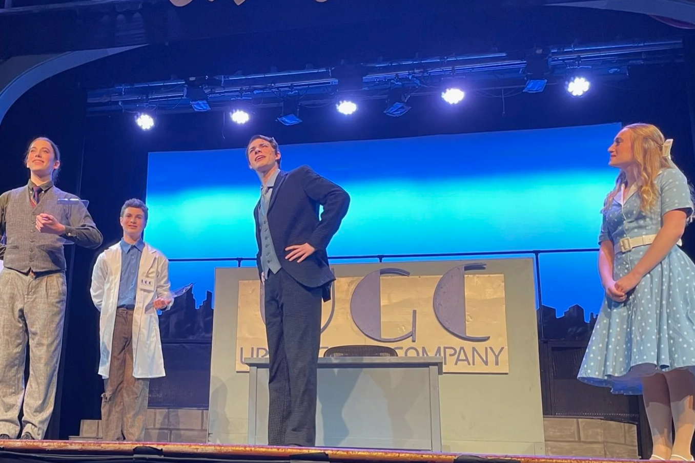 The cast of a theatrical play on stage with a blue background and a sign that reads 'UPG-UPD Company'. Four actors are visible, dressed in period costumes, standing with confident and happy expressions.