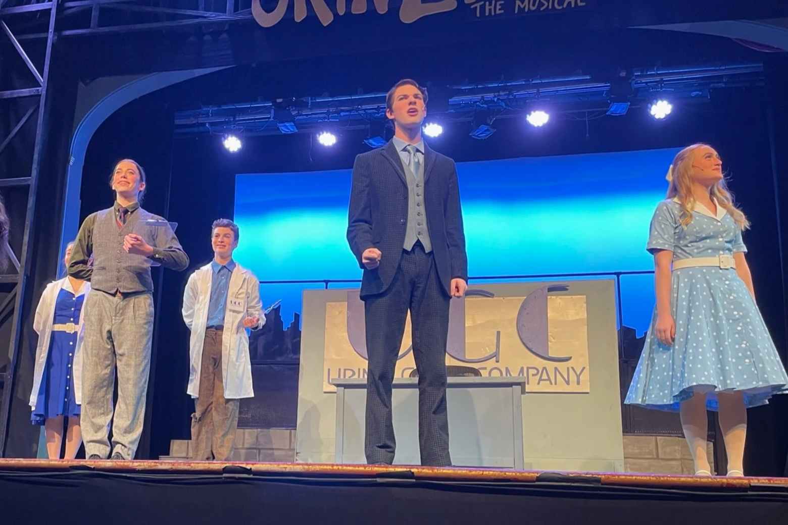 The cast of 'Once' musical on stage, dressed in vintage costumes, standing on stage with blue backdrop and a sign that reads 'Lippincott Company'.