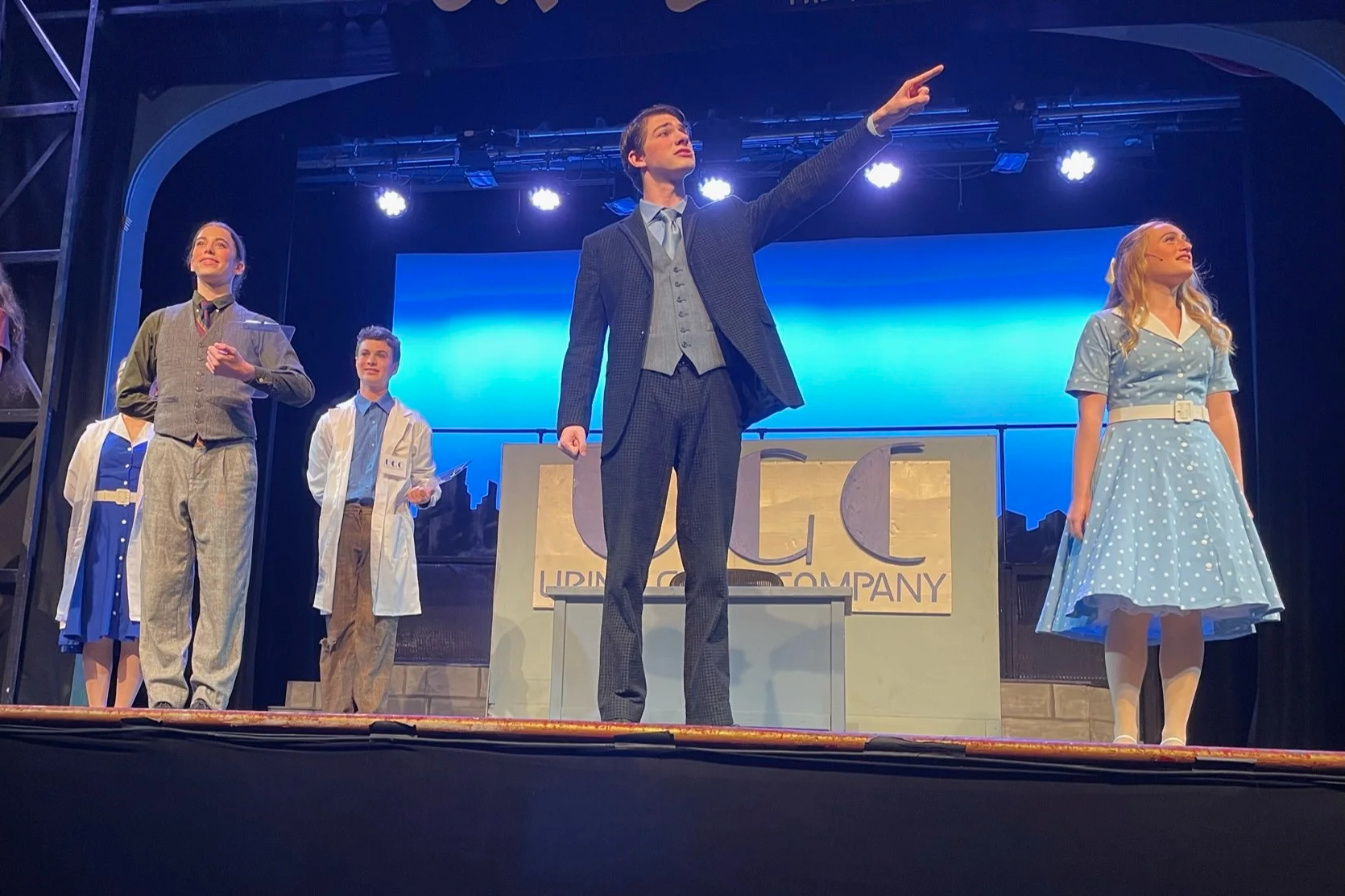 Group of five performers on stage during a theatrical production, with a blue background and stage lighting, dressed in vintage clothing.