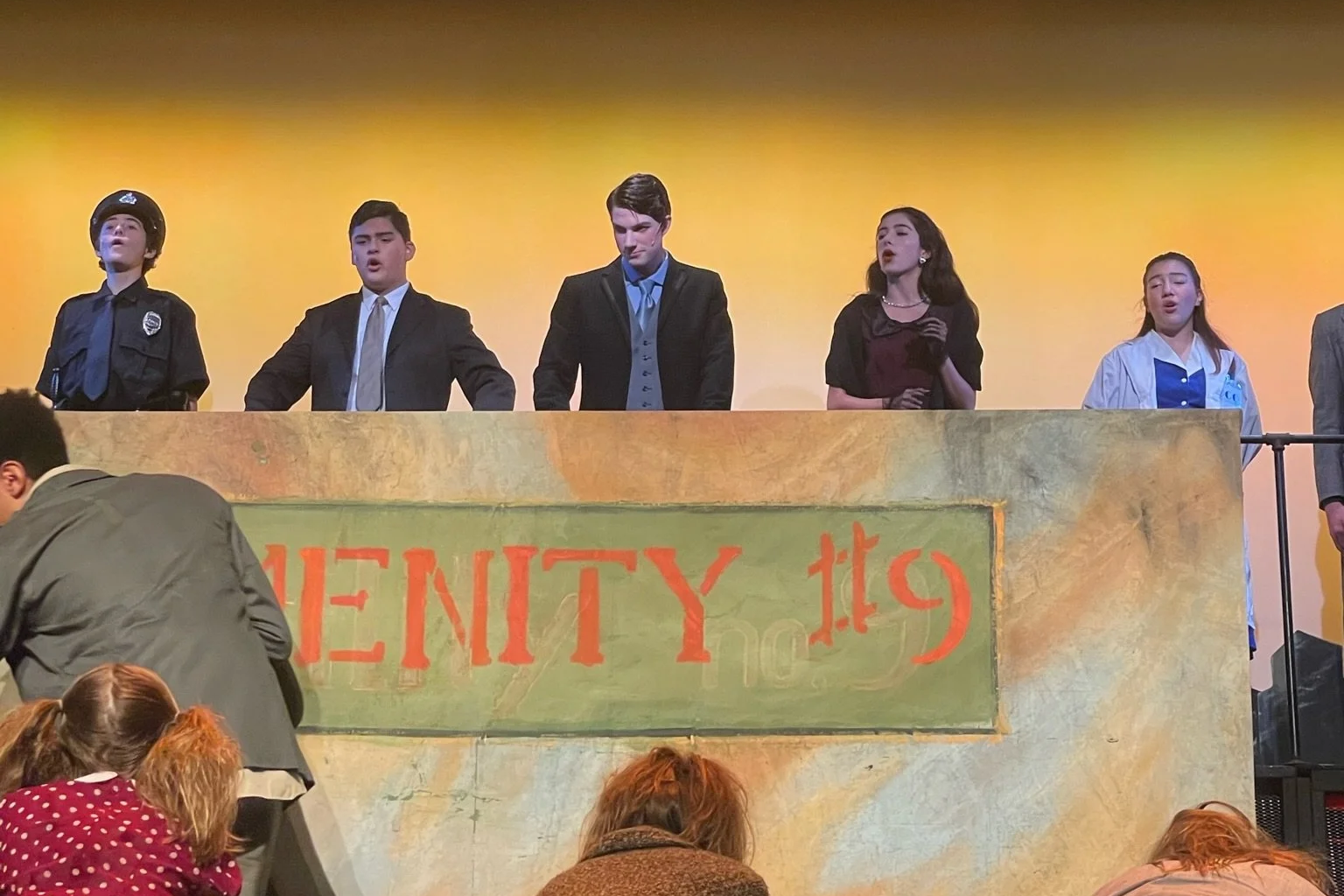 Group of young people performing on stage, with some dressed as a police officer, a professional, and a student, during a theatrical play or presentation.