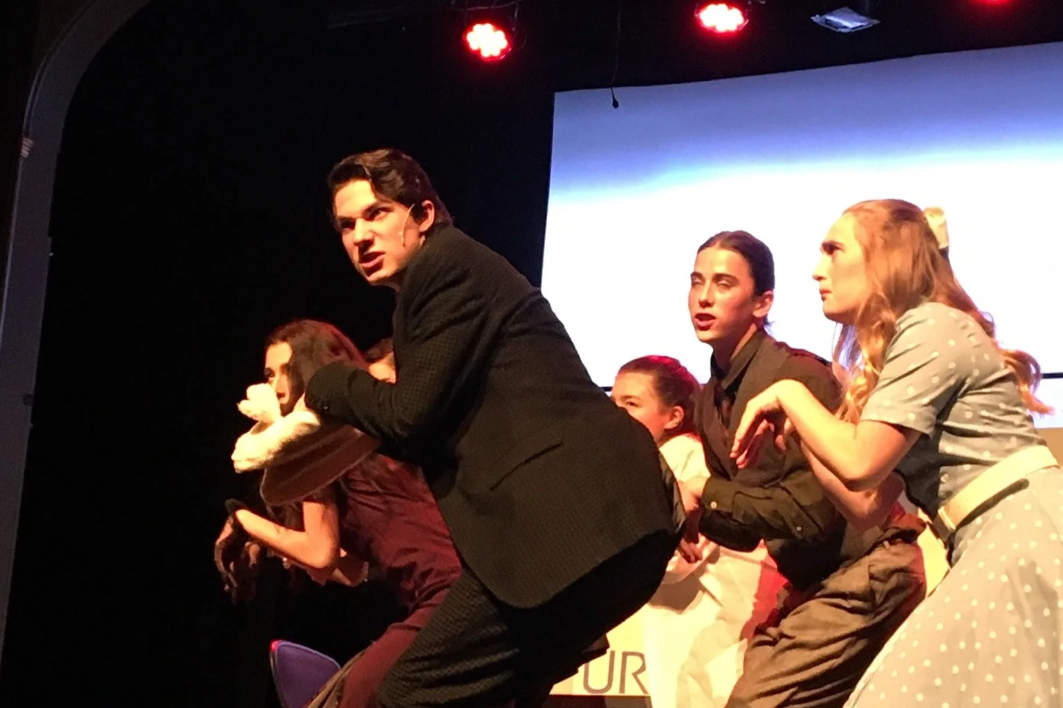 A theatrical group performing on stage, with a man in the foreground wearing a black suit, leaning forward and speaking, while others behind him, including women and children, react with various expressions.