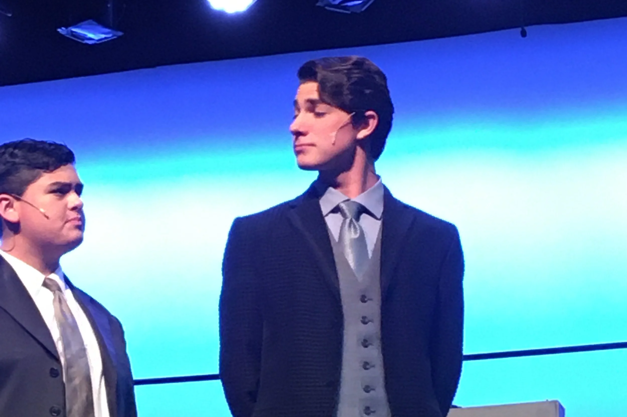 Two men in formal suits standing on stage, one with his eyes closed and the other looking at him, with a blue screen background.
