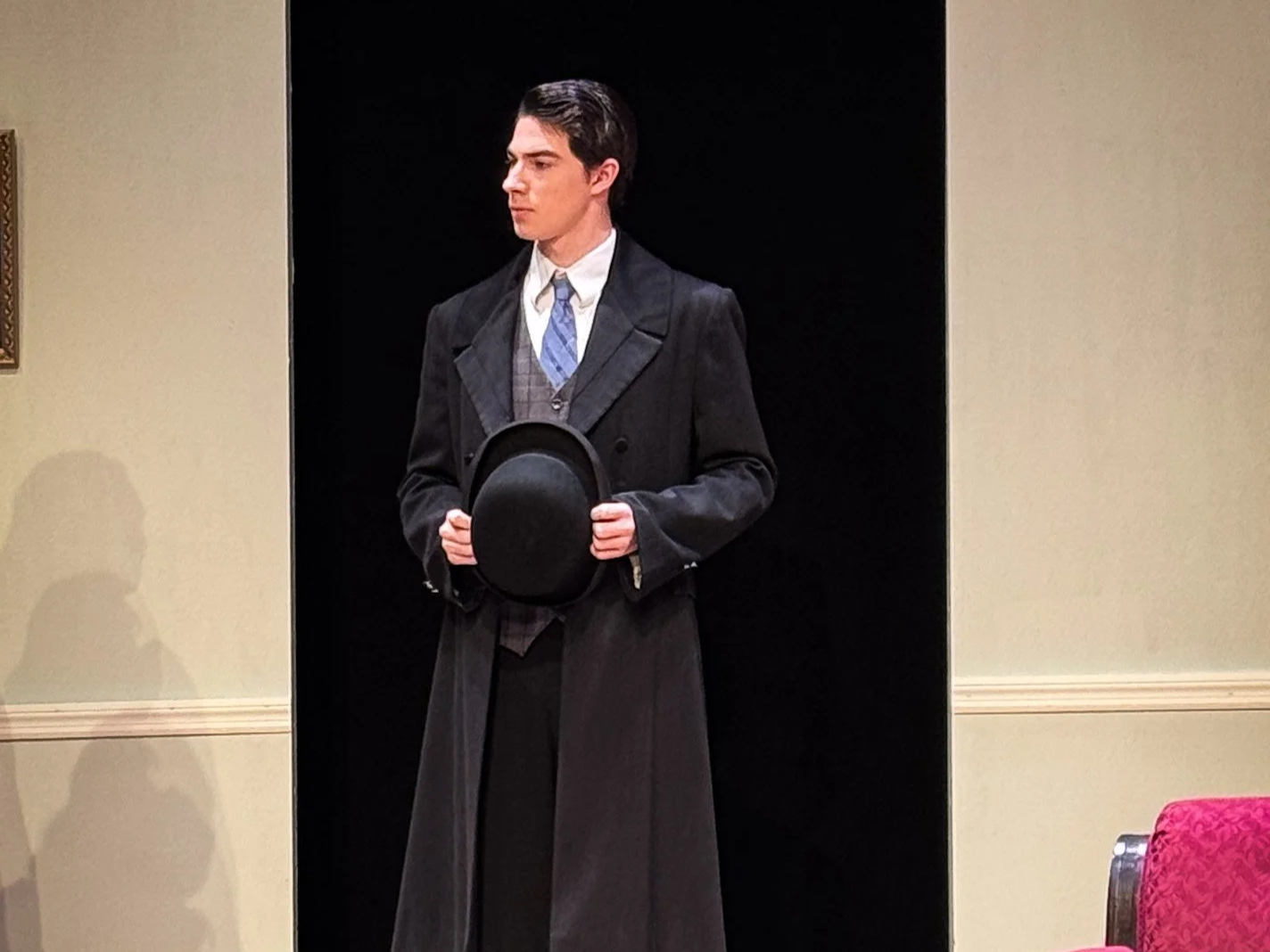 A young man dressed in a vintage black overcoat and gray vest, holding a black bowler hat, standing on stage with a dark background.