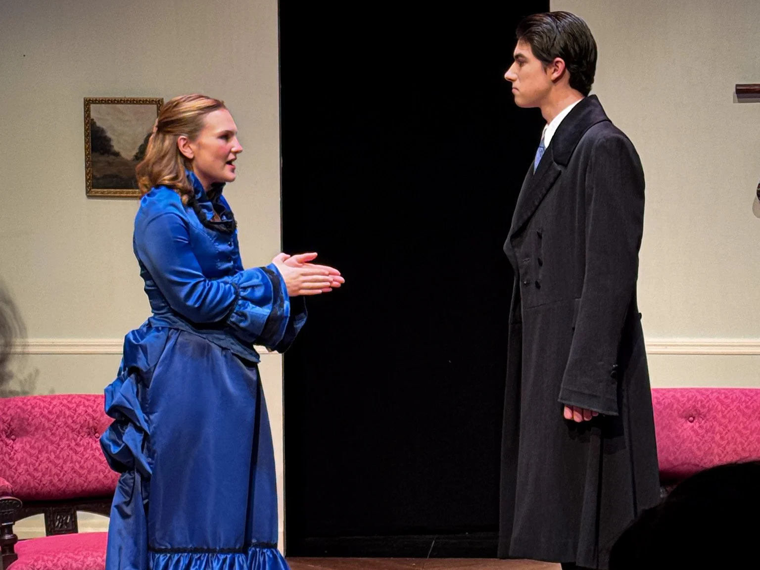 Two actors are performing a scene in a play. The woman on the left, dressed in a blue dress, is talking animatedly with her hands, while the man on the right, dressed in a black suit and long coat, is listening attentively.