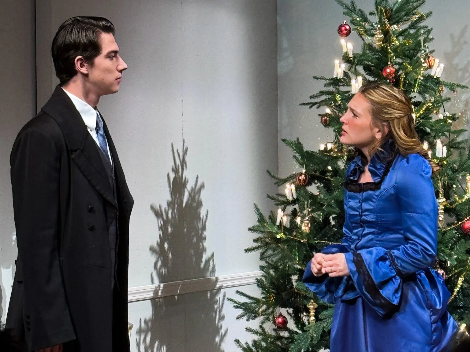 A man and a woman standing face-to-face near a decorated Christmas tree, indoors, with serious expressions.