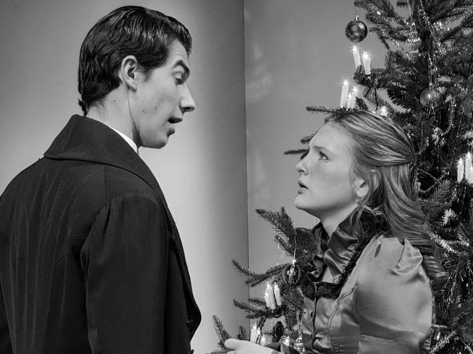 Black and white photo of a man and a woman standing face-to-face next to a decorated Christmas tree, looking at each other with serious expressions.