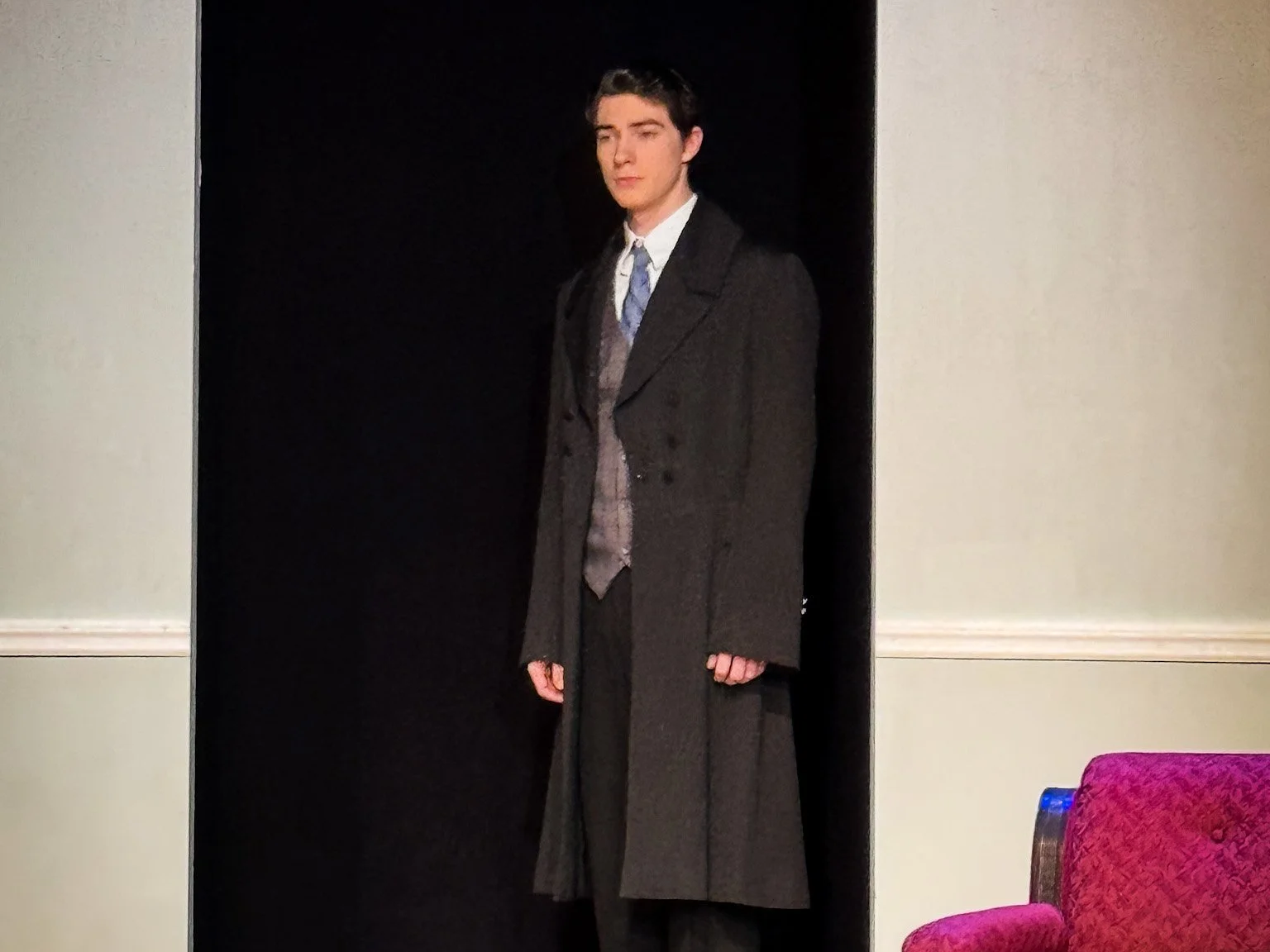 Young man in a formal suit and long coat standing in front of a black background, with cream-colored walls and a red upholstered chair visible in the foreground.
