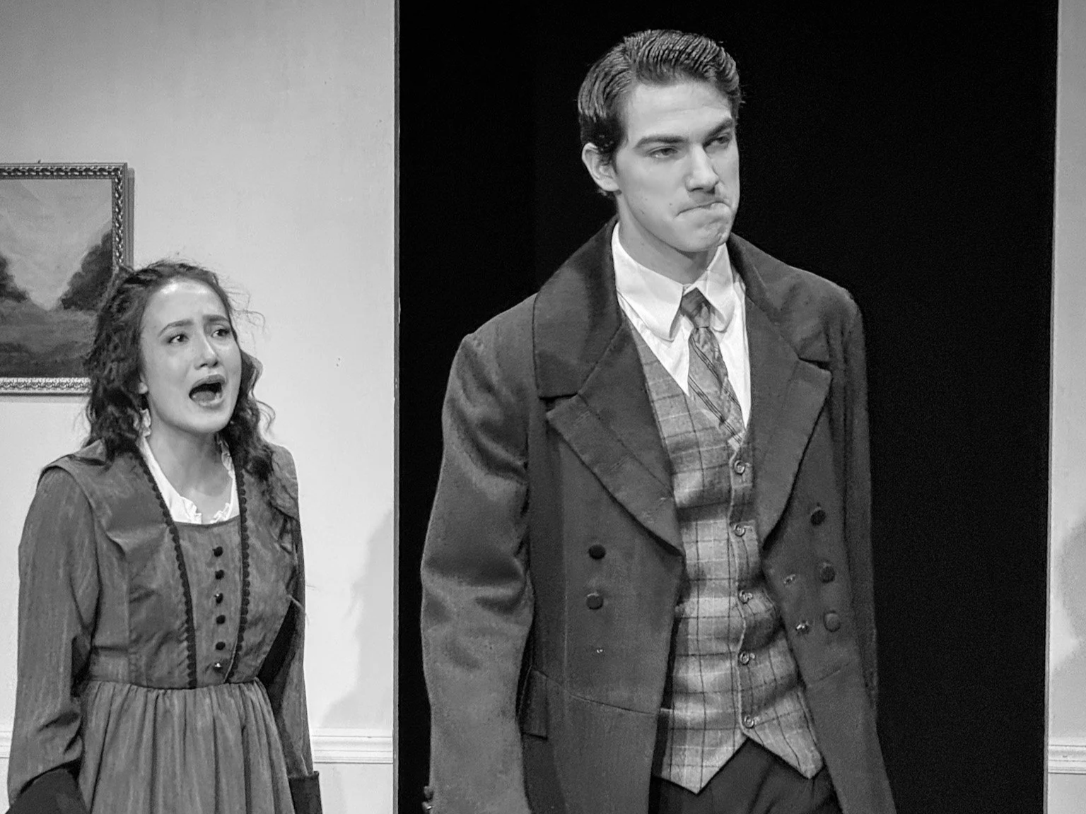 A black-and-white scene from a theatrical play featuring a woman with curly hair, wearing a vintage dress, appearing distressed, and a young man with neatly combed hair, dressed in a period suit and coat, standing with a serious expression