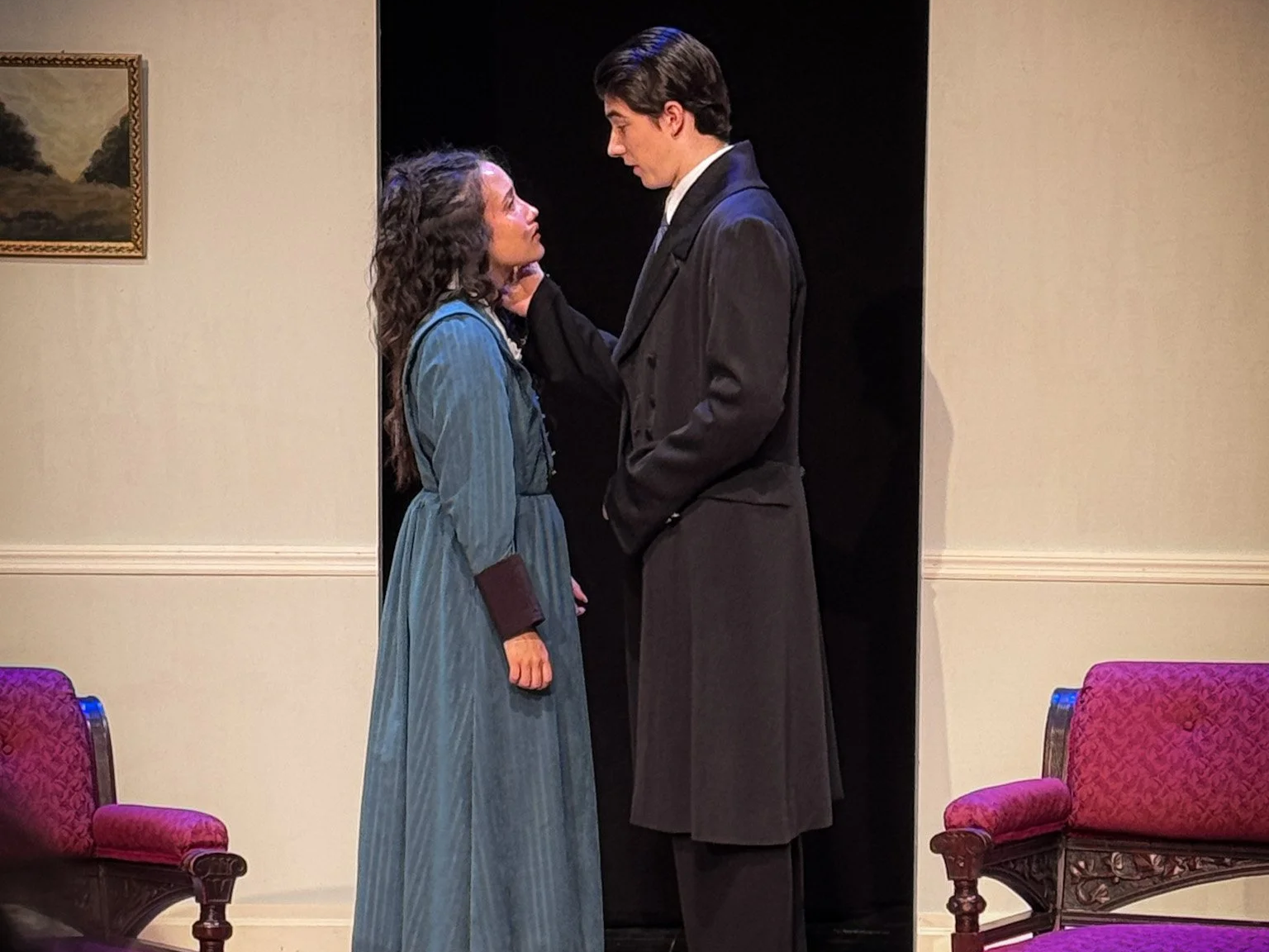 Two people, a woman and a man, stand close together in a room, looking into each other's eyes. The woman is wearing a dark blue dress and the man is wearing a dark suit. They are centered in the image, with purple chairs on either side. The setting a