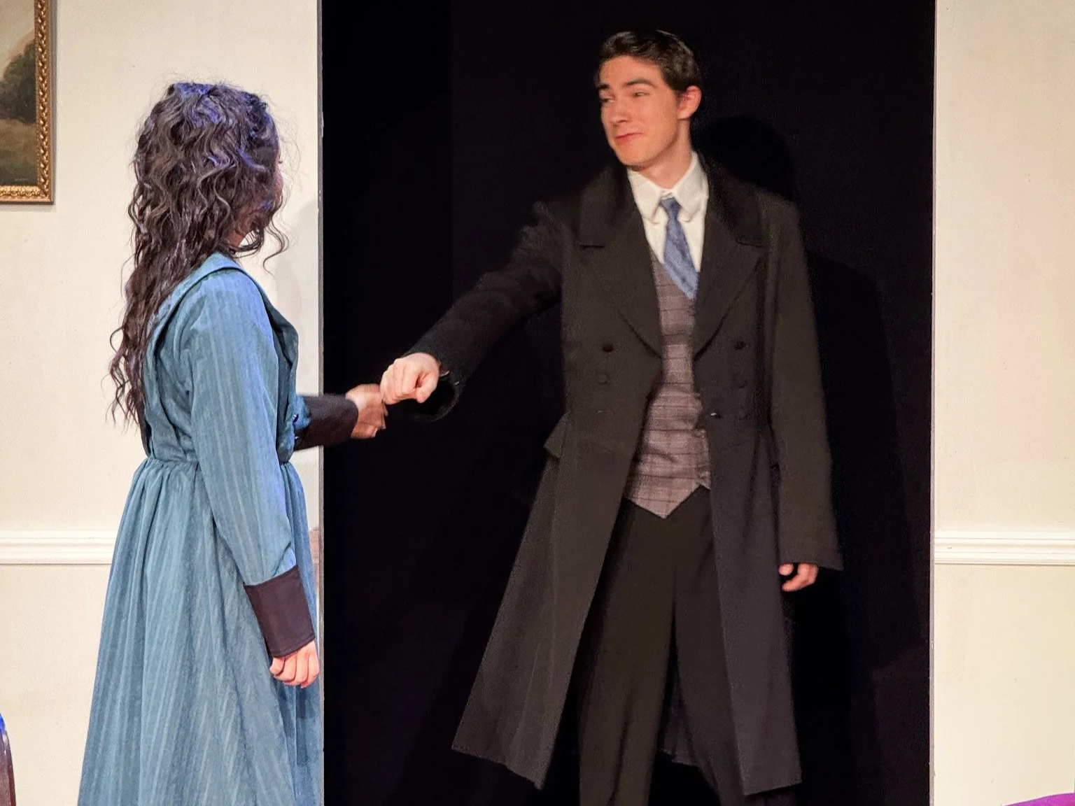 A woman in a blue dress shaking hands with a man in a long coat standing in front of a black doorway.