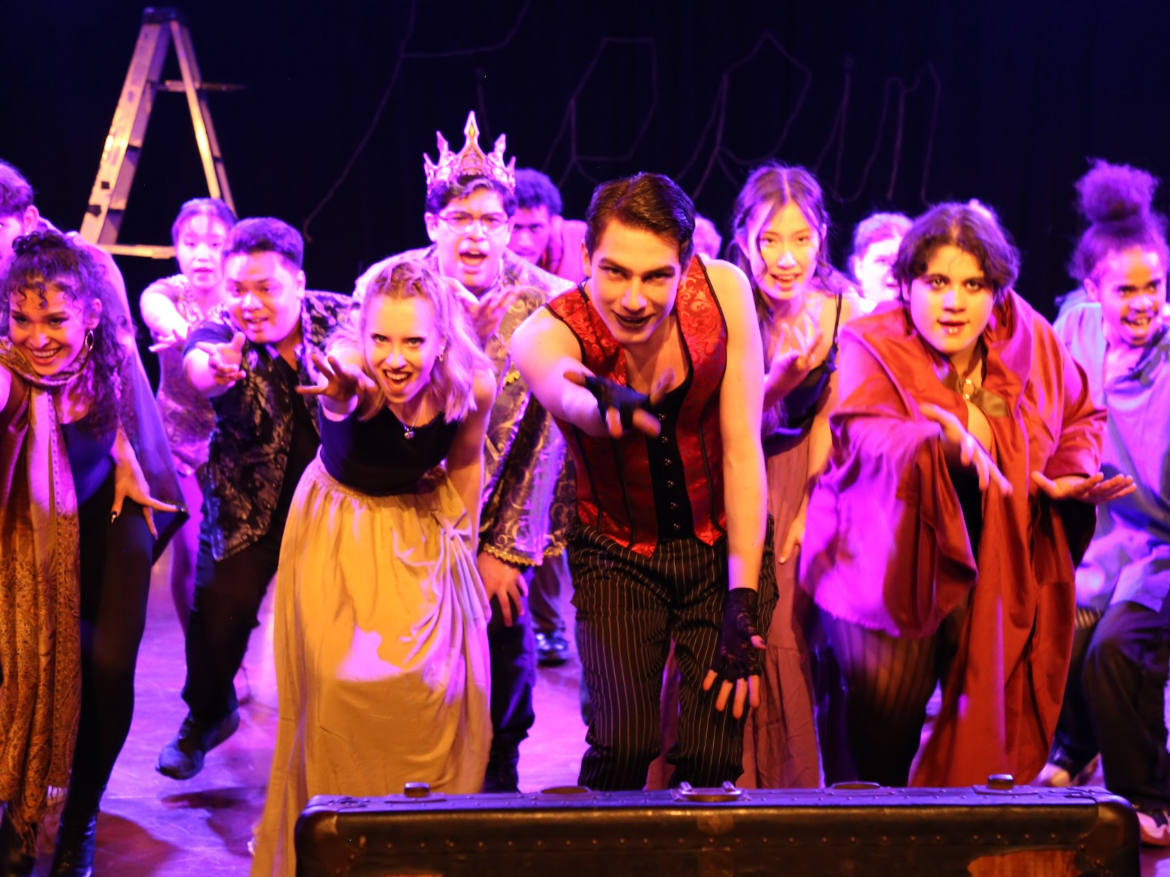 Theatrical cast performing a lively scene on stage with purple lighting, featuring a man in a corset and striped pants, and a group of actors in colorful costumes.