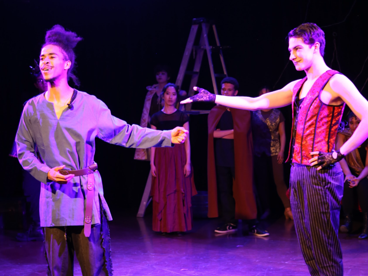 Two performers on stage with others observing in the background. One performer is extending an arm toward the other. The scene is illuminated with purple lighting, and stage equipment like a ladder is visible.
